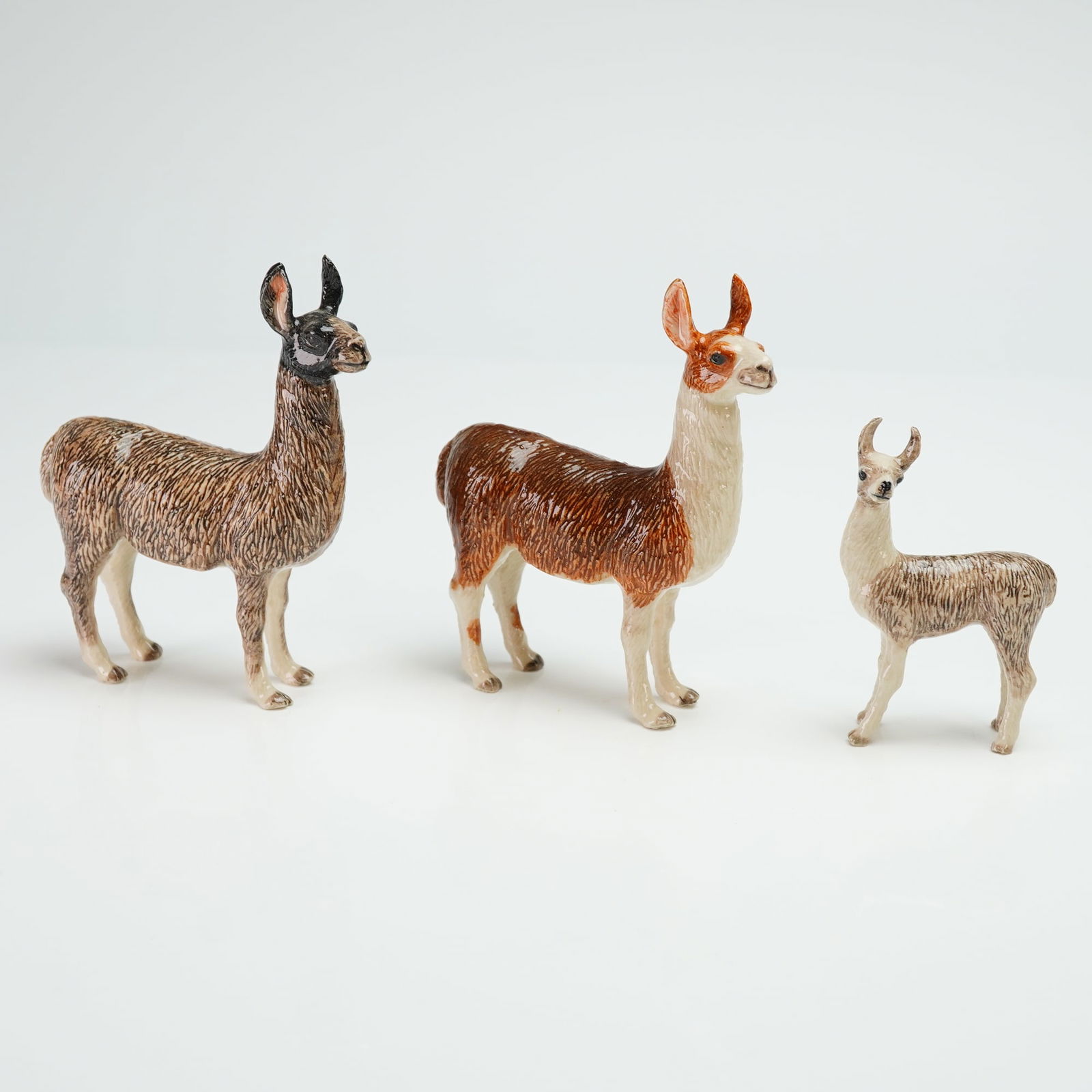 Three Glazed Porcelain Llama Figurines: Figurines featuring naturalistically modeled forms with textured surfaces representing wool. The collection consists of three hand-painted figures in varying sizes and colorations. The largest figure