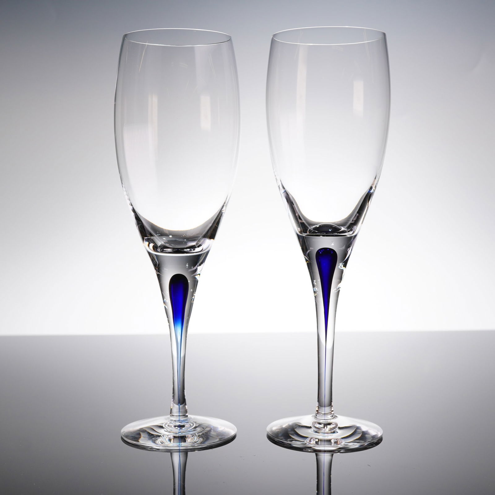 A Pair of Orrefors Intermezzo Blue Champagne Flutes: A pair of contemporary stemware pieces identified as Orrefors Intermezzo Blue. The set consists of two champagne fi?utes, constructed from clear crystal. Each fi?ute features a tall, slender stem