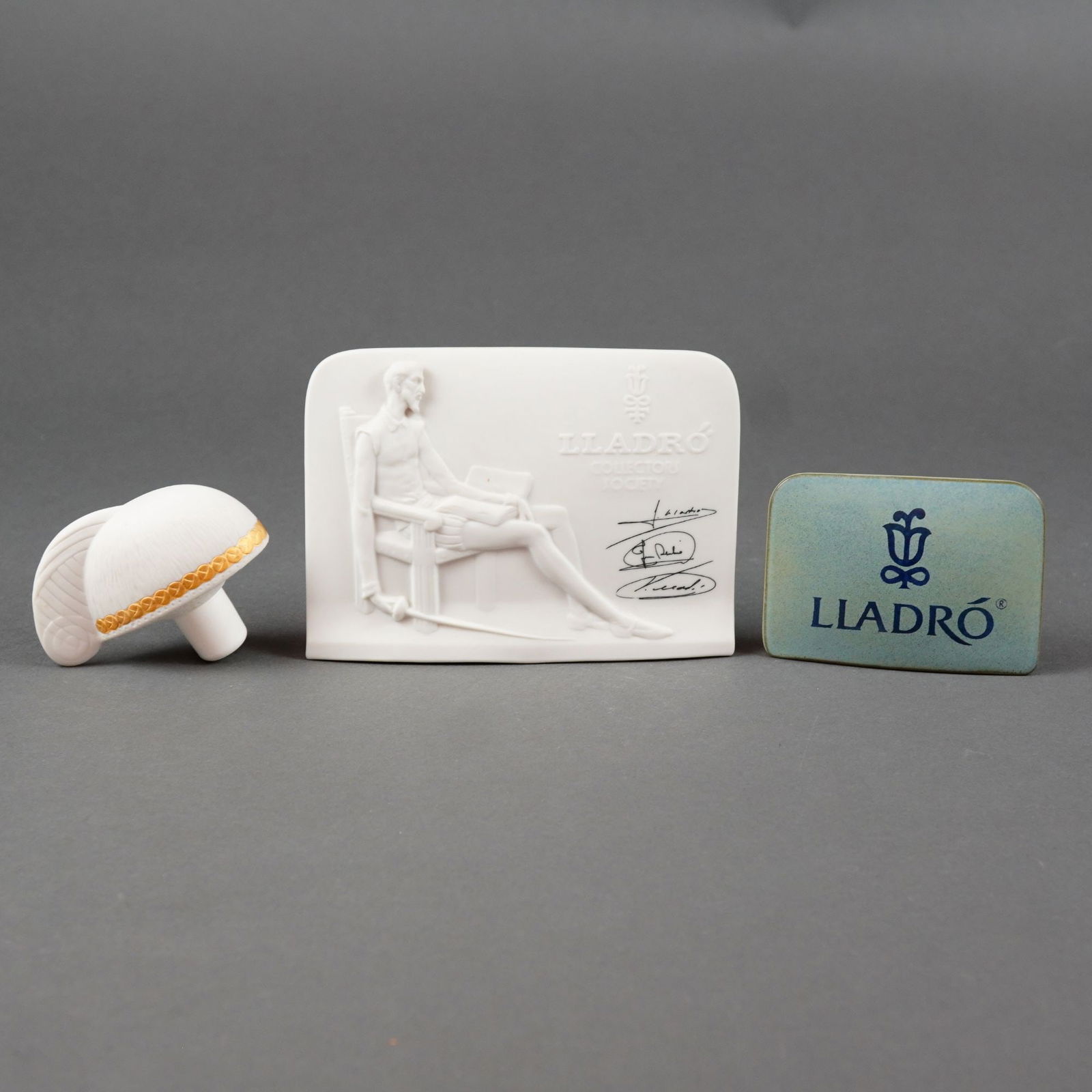 Lladro Collectors Society Signs and Centurion Hall Bottle Stopper: A three-piece Lladro grouping featuring two signs and a Centurion Hall bottle stopper. The set includes a 1985 Lladro Collectors Society display plaque in white bisque porcelain, featuring a low-relie