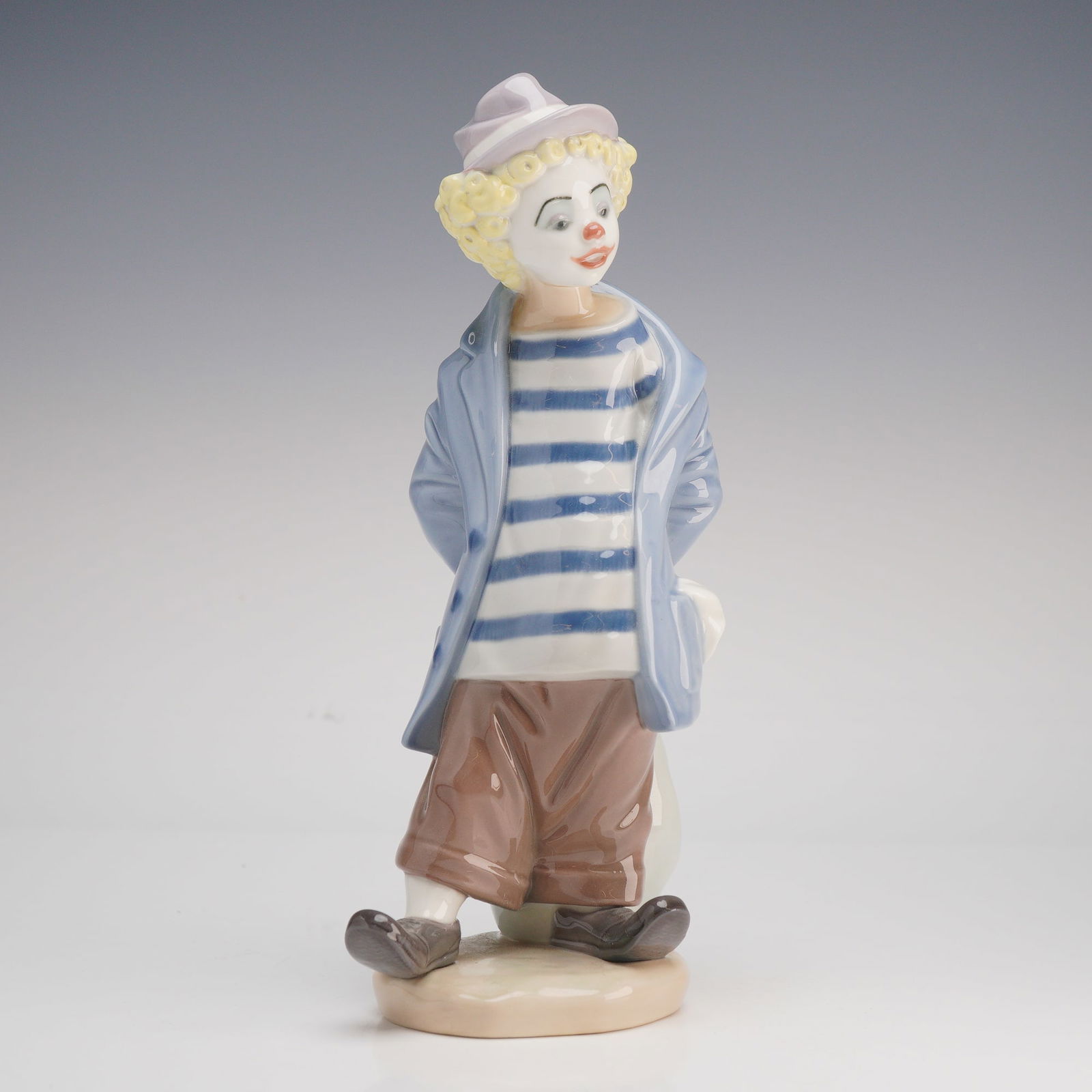 Lladro Little Traveler Porcelain Figurine Number 7602: A Lladro glazed porcelain figurine titled Little Traveler, model number 7602. This piece depicts a young clown standing in a light blue overcoat over a blue and white striped shirt and brown