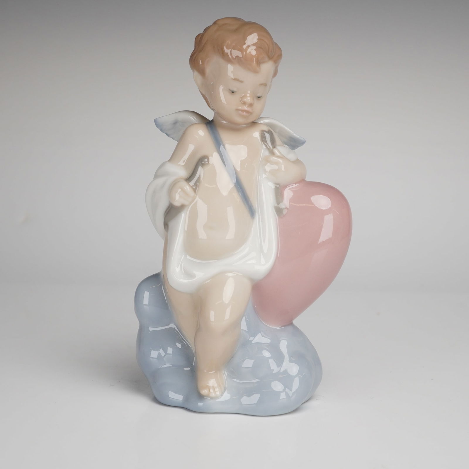 NAO by Lladro Porcelain Figurine, Cupid: A NAO by Lladro glazed porcelain figurine titled Cupid, model number 1436. The figure depicts a winged Cupid standing upon a pale blue cloud-form base and leaning against a large pink heart. The