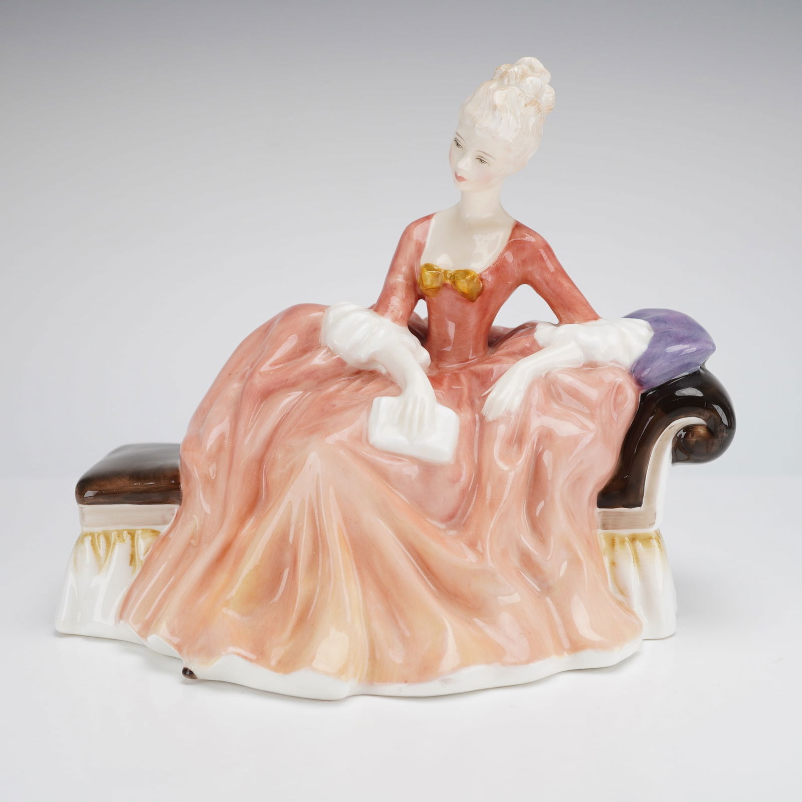 Royal Doulton Figurine Reverie HN2306: A glazed bone china figurine by Royal Doulton, depicting a woman in a pinkish-orange dress seated on a brown and white chaise. Marked on the base with the Royal Doulton crown mark, name, and model