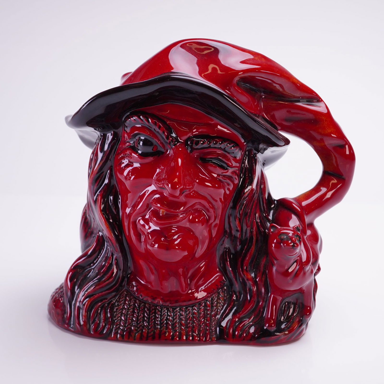 Royal Doulton Flambe Large Character Jug, The Witch D7239: Finished in a signature high-gloss red and black flambe glaze. The ceramic piece features a stylized bust of a witch with a cat on her shoulder and a pointed hat that curves to form the handle. The ba