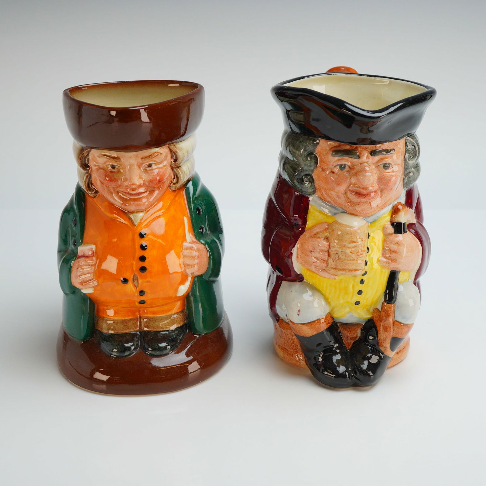 Two Royal Doulton Character Toby Jugs- D6319 and D6109: Two-character jugs comprise this lot including The Squire D6319 and Jolly Toby D6109. The Squire depicts a seated figure in an 18th-century style green coat and orange waistcoat holding a long pipe an