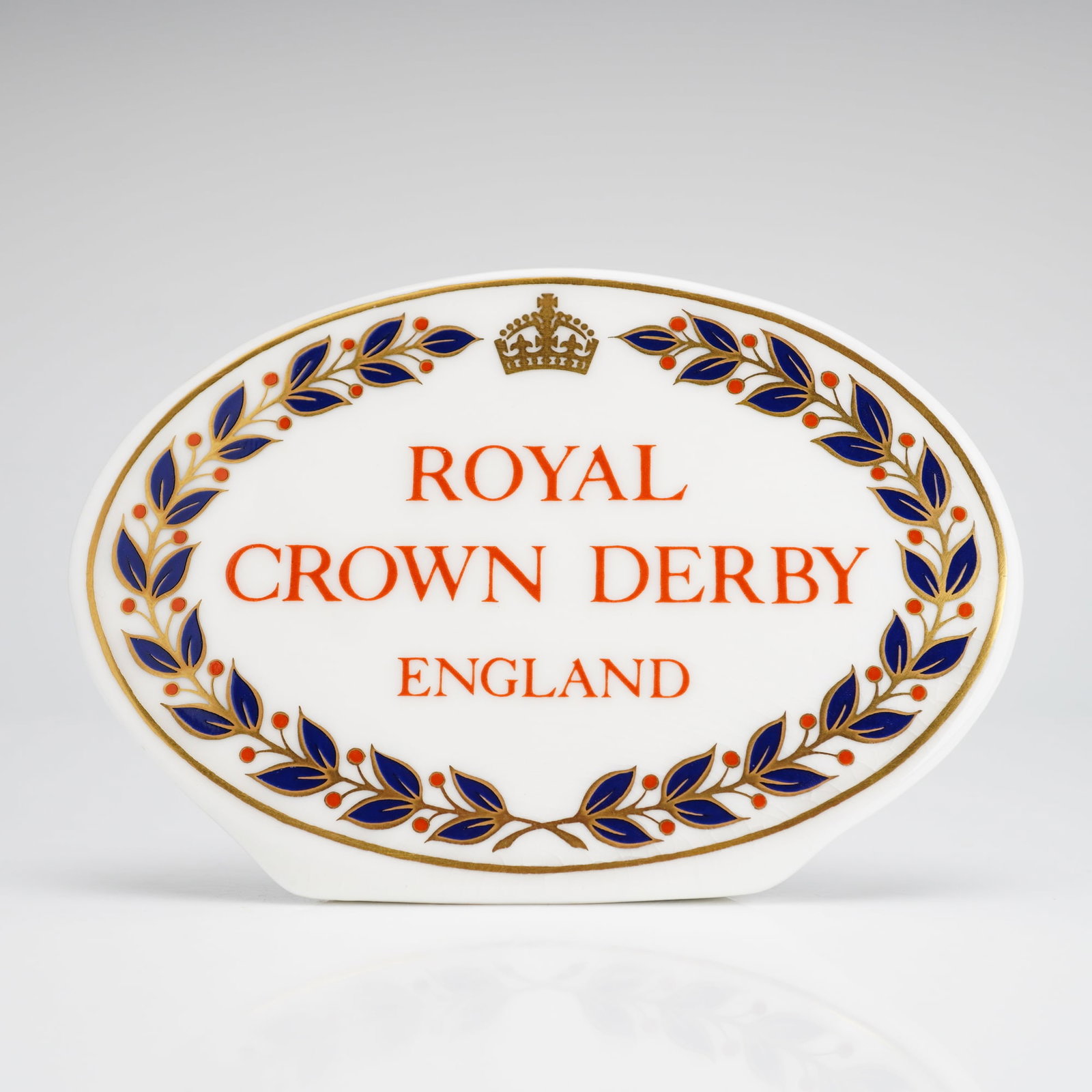Royal Crown Derby Dealer Display Plaque: A Royal Crown Derby English bone china name plaque designed for use as a retail dealer display sign. The piece is crafted from glazed white bone china in a self-standing, curved oval form. The face fe