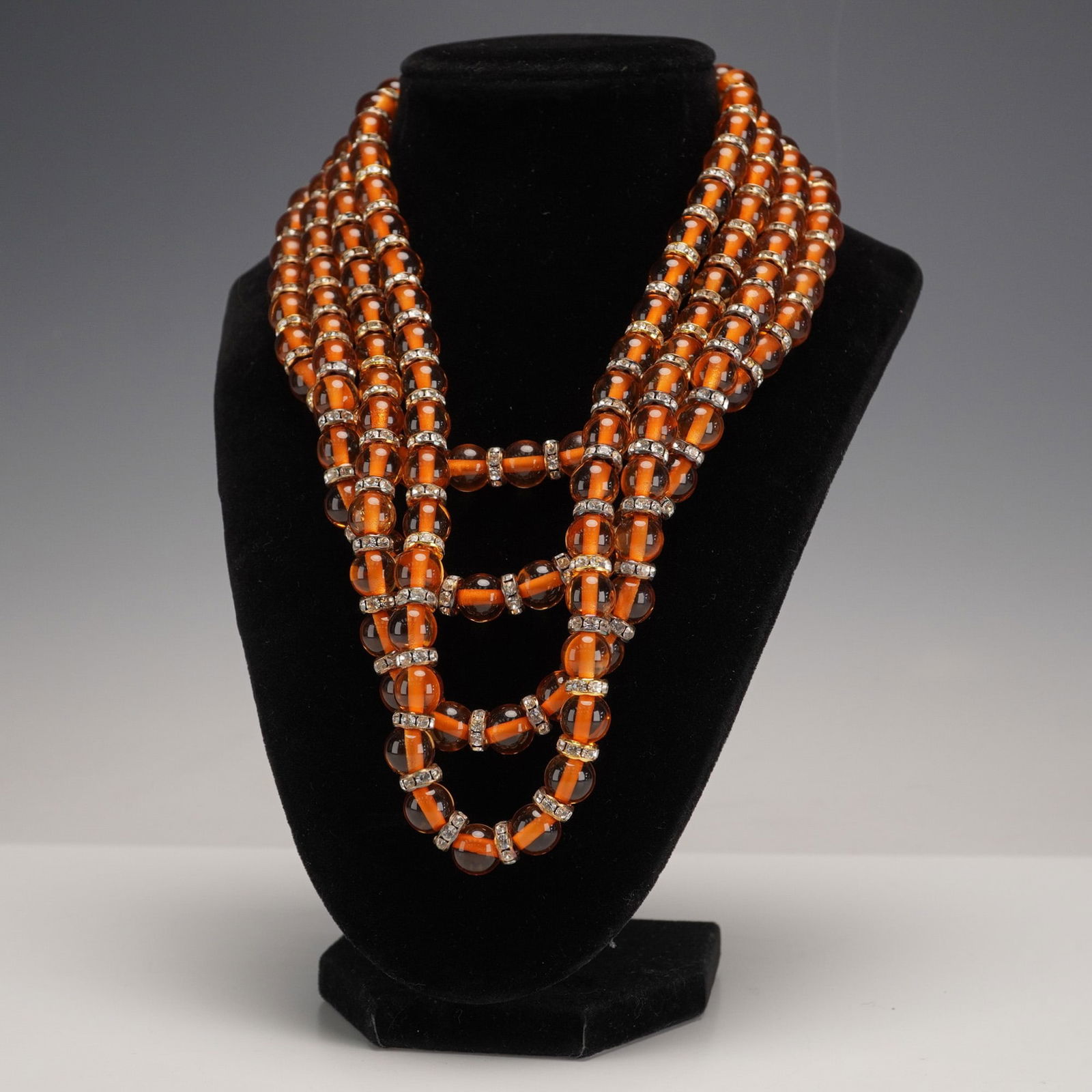 Valentino Four-Strand Amber Glass Bead Necklace with Rhinestone: Crafted with transparent amber-orange glass or acrylic beads, each featuring an opaque orange core or strip visible within. Interspersed throughout the strands are gold-toned metal rings, each set wit
