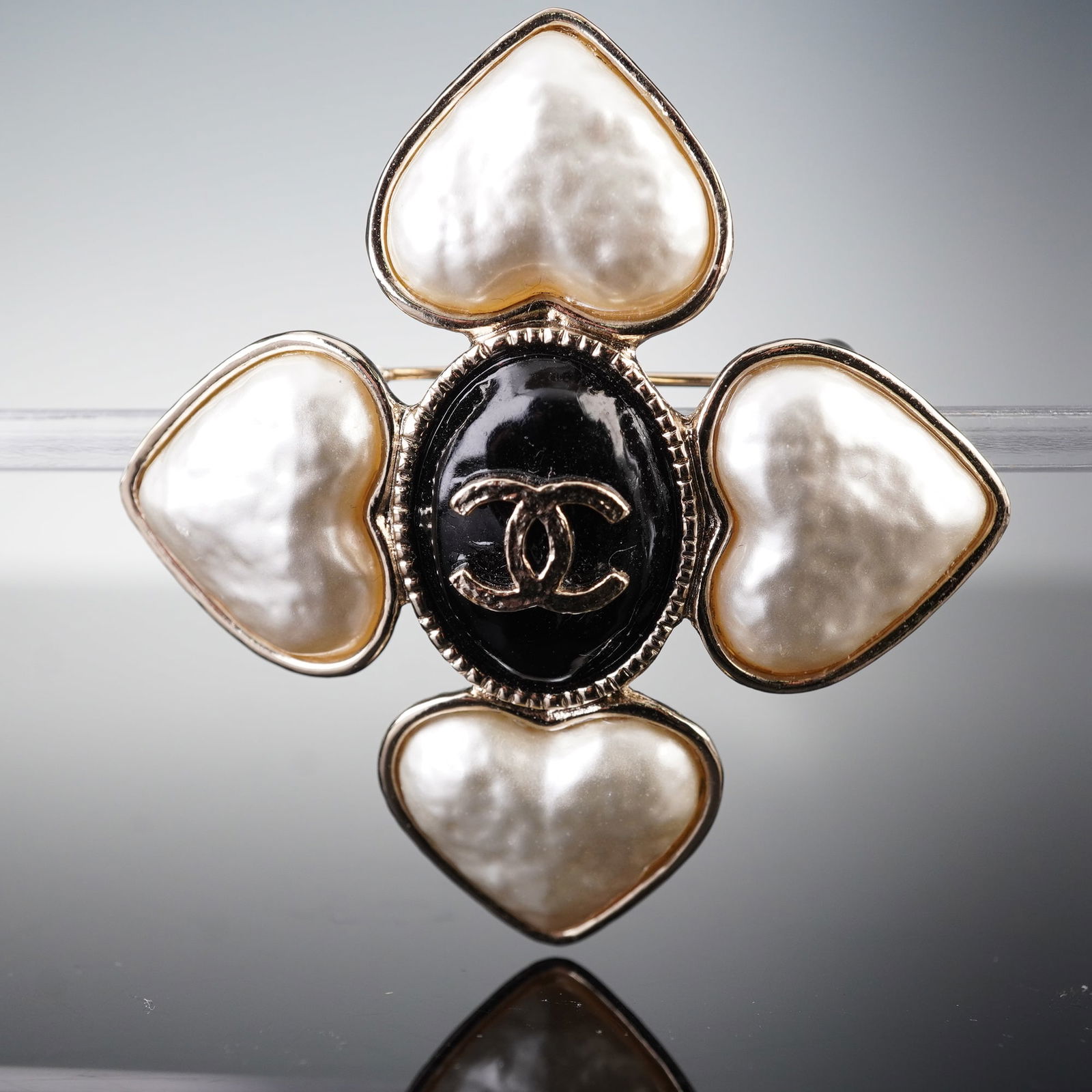 CHANEL B22 Hearts CC Brooch: A CHANEL B22 Hearts CC Brooch, crafted as gold-tone costume jewelry. The brooch features a central black oval element with an embossed gold-tone interlocking 'CC' logo, surrounded by four prominent he