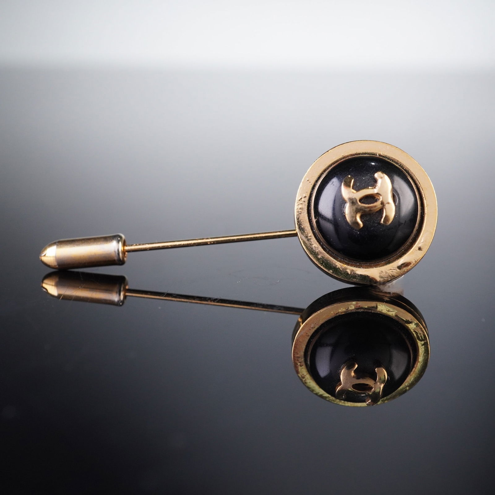 A CHANEL CC Logo Gold-Tone Stick Pin: A CHANEL CC Logo stick pin or lapel pin, featuring a circular head with a central black inlay prominently displaying a stylized gold-tone interlocking 'CC' motif, all encircled by a gold-tone bezel. T