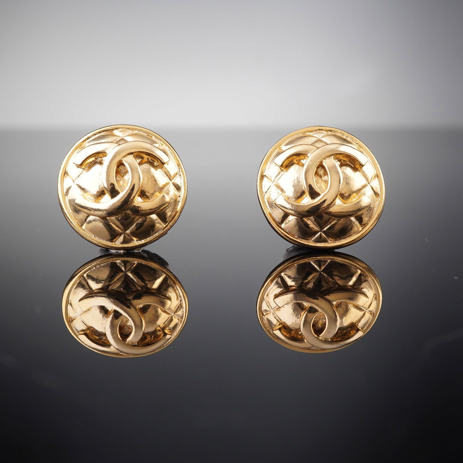 A Pair of Vintage Chanel Gold-Tone Quilted Matelasse Buttons: A pair of vintage Chanel gold-tone quilted matelasse buttons. Each circular, domed earring features a prominent interlocking 'CC' logo at its center, surrounded by a raised quilted or matelasse patter