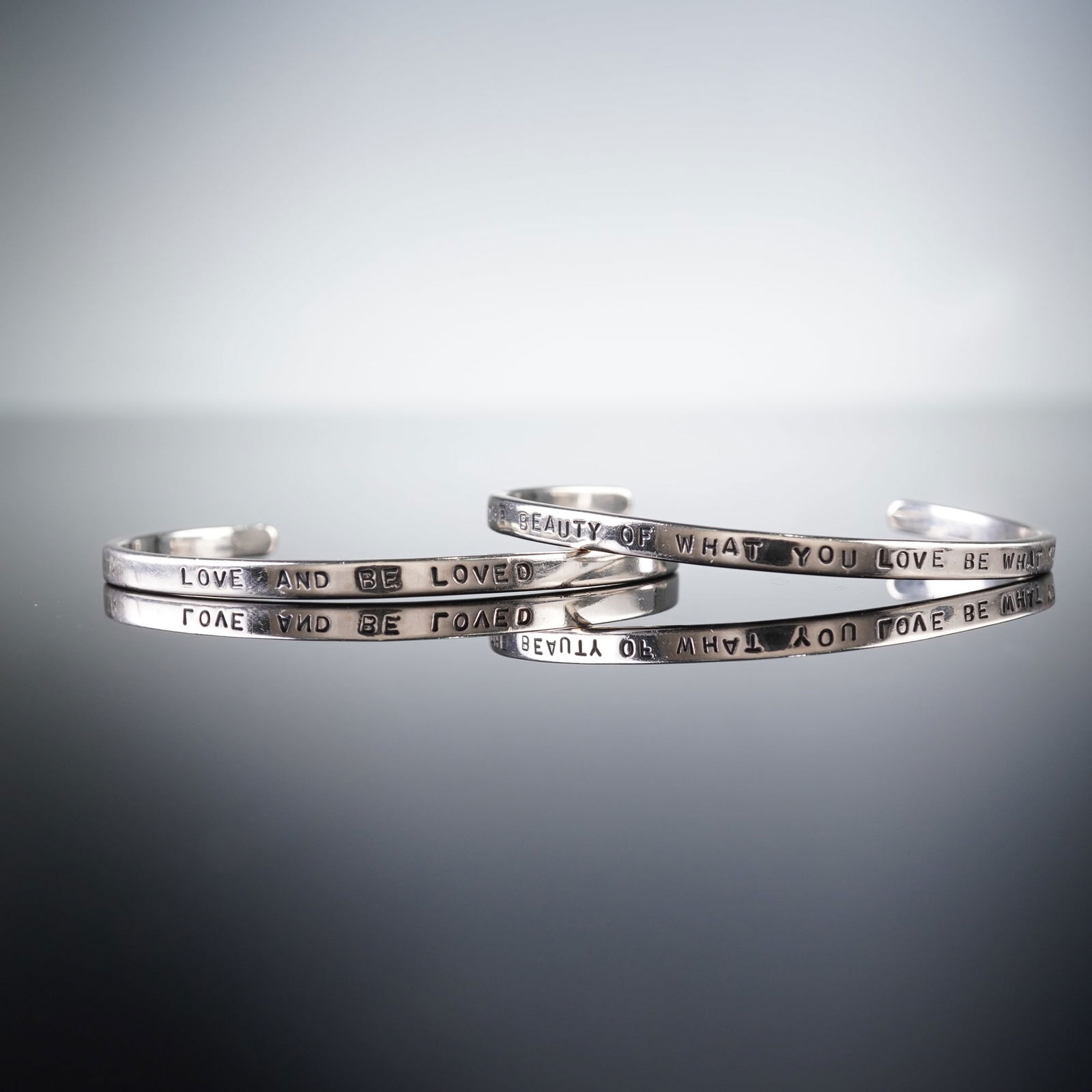 A Pair of Sterling Silver Mantra Cuff Bracelets: A pair of contemporary sterling silver Mantra Cuffs. The minimalist, open-band design features deeply stamped text in capital letters. The first bracelet is inscribed "LOVE AND BE LOVED," and the seco