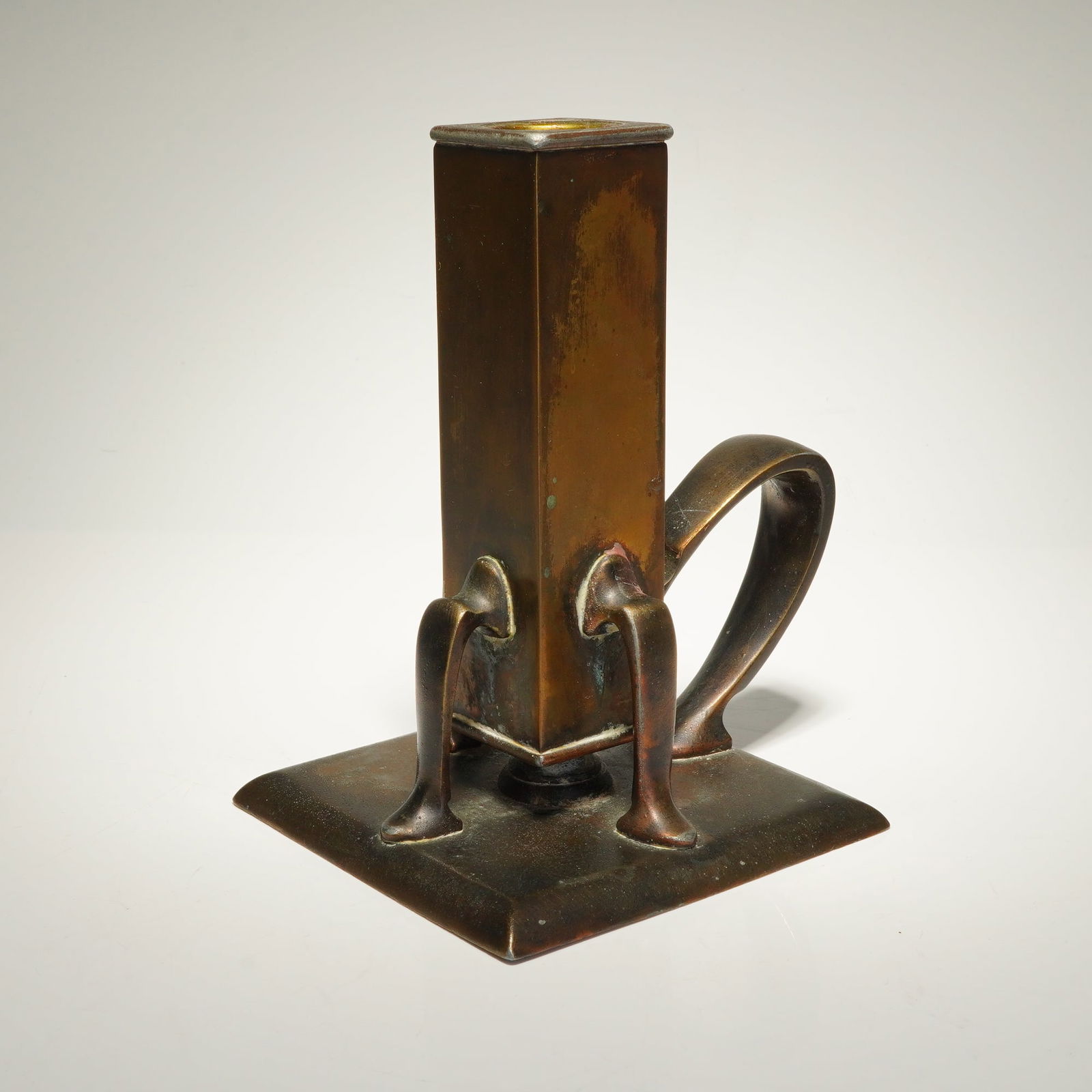 Brass Square Chamberstick Candle Holder: A brass chamberstick featuring a deep, aged brown patina. The form consists of a tall, square columnar candle socket with a circular opening at the top. The socket is supported by three stylized curve