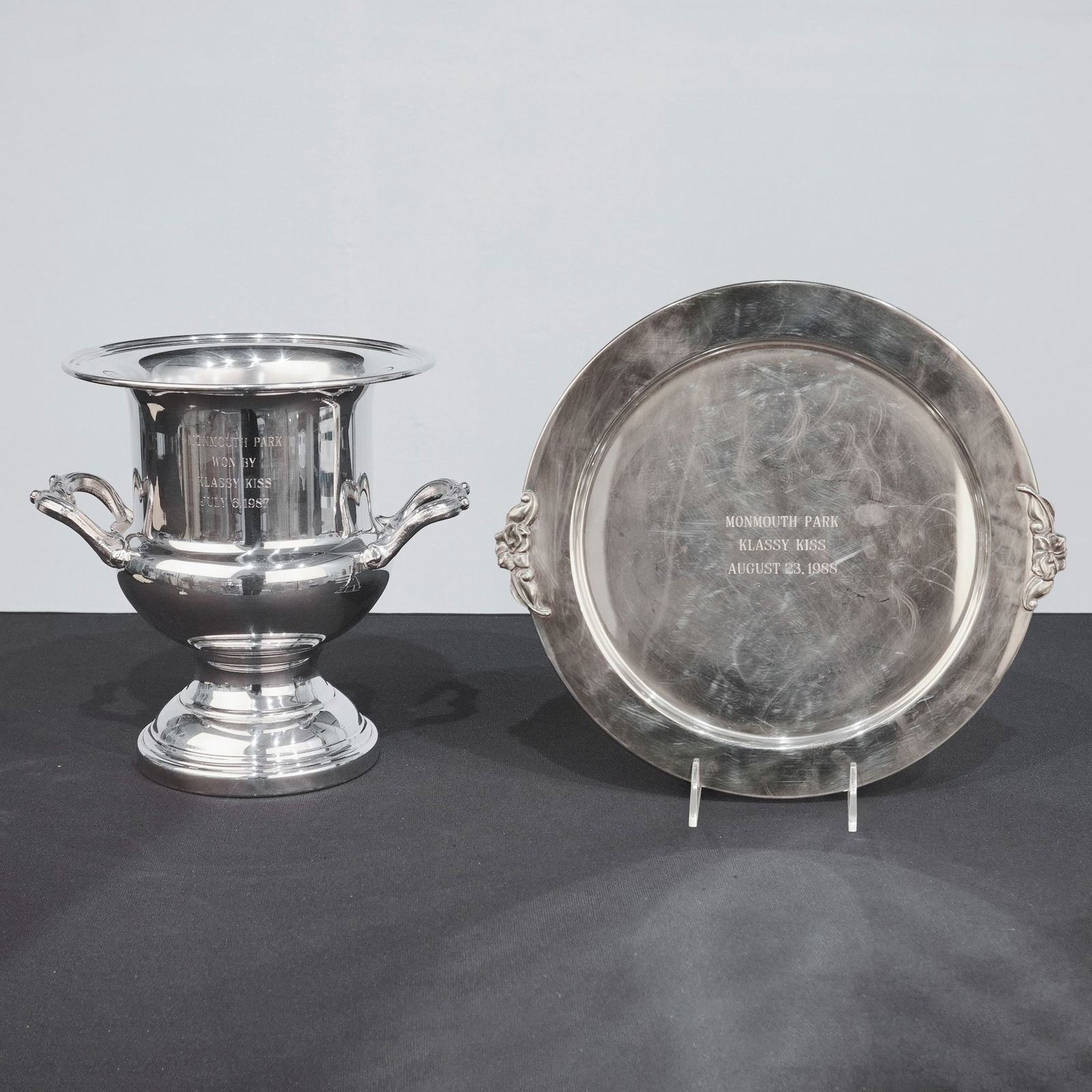 2pc Monmouth Park Silver-Plate Horse Racing Trophies Awarded to Klassy Kiss: A two-piece collection of silver-plated equestrian trophies awarded at Monmouth Park to the horse Klassy Kiss in the late 1980s. The group includes a two-handled pedestal trophy bucket manufactured by