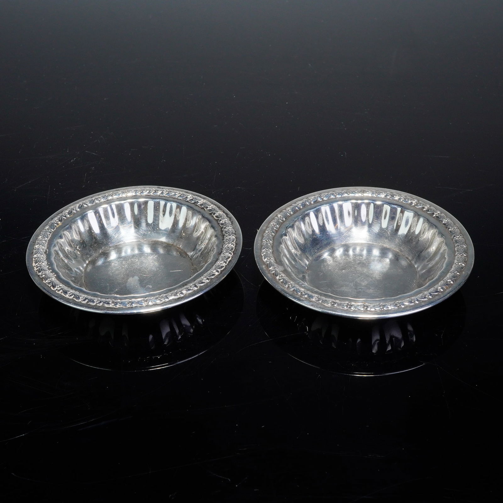 Pair of Sterling Silver Small Round Dishes: A pair of sterling silver small round dishes. Each dish is designed with a circular form featuring a fluted well and an ornate embossed foliate border. Stamped on the base with hallmark and STERLING.