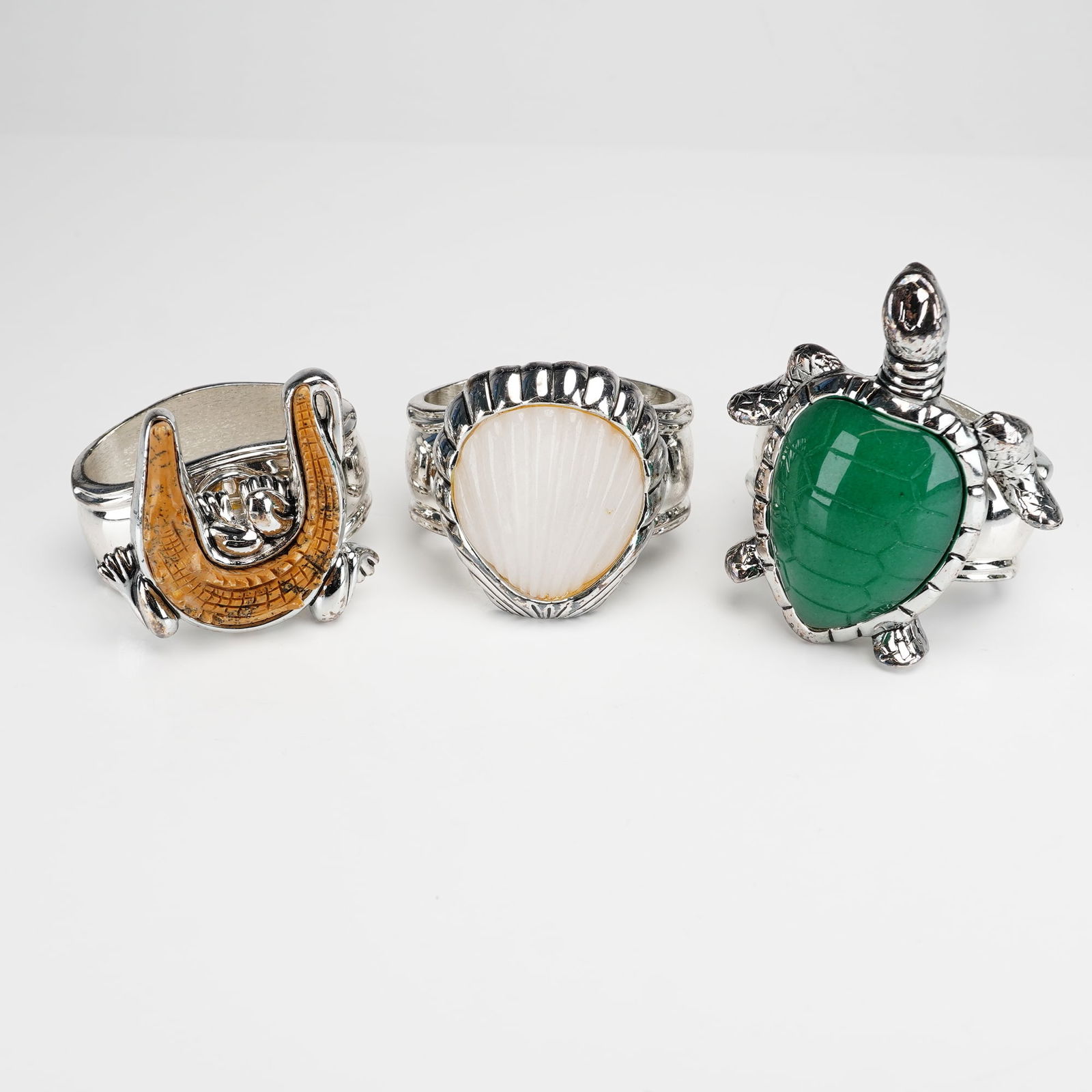 Three Hans Turnwald Silverplated Napkin Rings: The collection features three distinct figurative motifs: a sea turtle with an inset polished green stone-like shell, a ribbed white shell-form inlay, and a carved ochre-colored alligator. Each ring i
