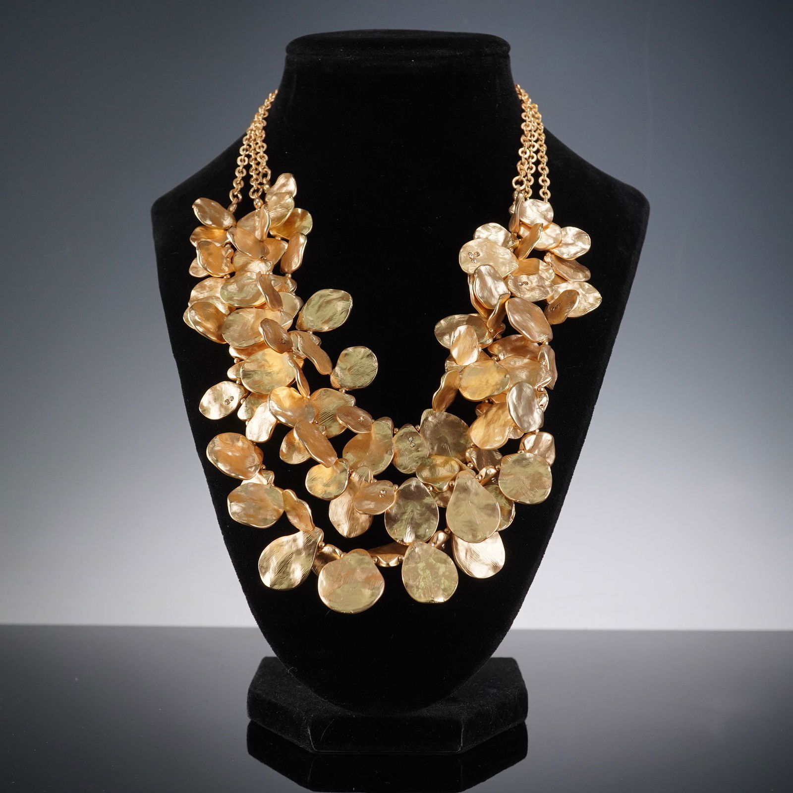 Gold-Tone Metal Hydrangea-Form Bib Necklace: A gold-tone metal bib-style necklace, featuring a dense, multi-layered arrangement of numerous hammered or textured leaf-like elements forming a hydrangea-form design. It is suspended from a double-st