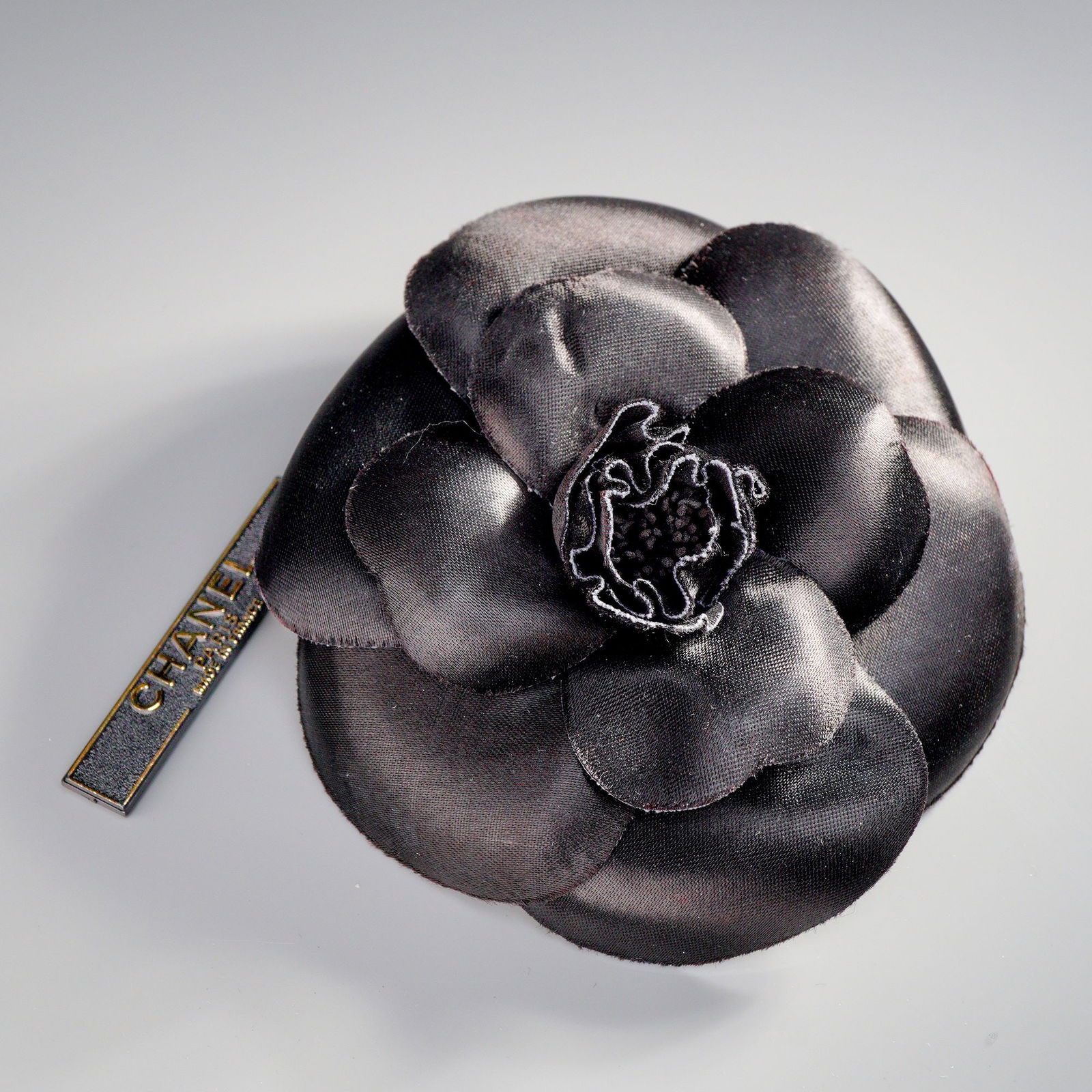 Chanel Paris Black Fabric Camellia Flower Brooch with Presentation Box: A Chanel black Fabric camellia flower brooch constructed from layered black woven satin, forming the characteristic camellia flower design. The reverse features a safety pin closure mechanism. Attache