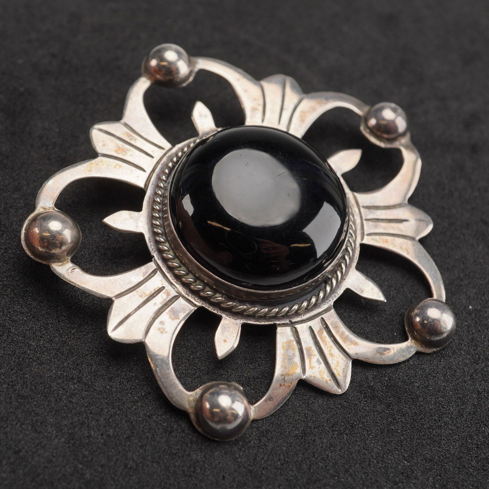 Mexican Sterling Silver Black Onyx Cabochon Openwork Brooch: A sterling silver brooch featuring a stylized openwork design. The center is dominated by a large, domed, circular black onyx cabochon, set within a twisted wire rope bezel. The outer framework is