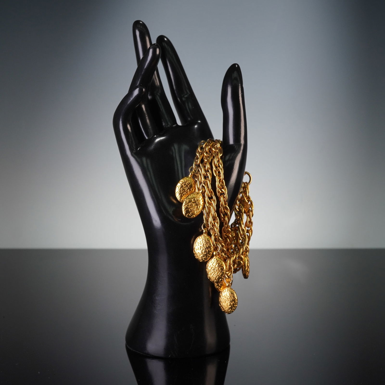 CHANEL Gold Tone Multistrand Charm Bracelet: A CHANEL gold-toned metal multistrand charm bracelet. The bracelet features multiple intertwined chain links adorned with numerous oval-shaped charms, each embossed with the repeated text CHANEL. It s