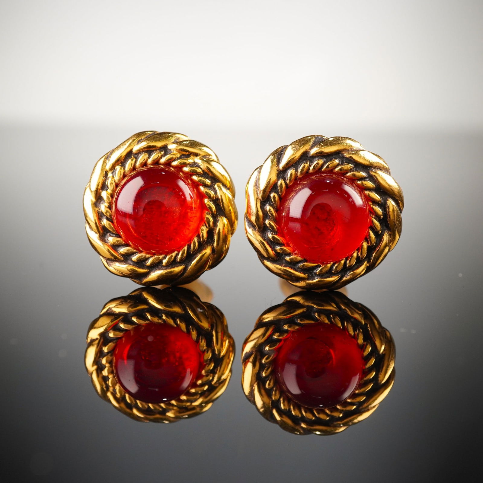 Chanel Gripoix Red Cabochon Earrings: A pair of Chanel Gripoix earrings, each featuring a central domed, translucent red glass cabochon-style element set within a substantial gold-tone metal bezel. The bezel is characterized by a prominen