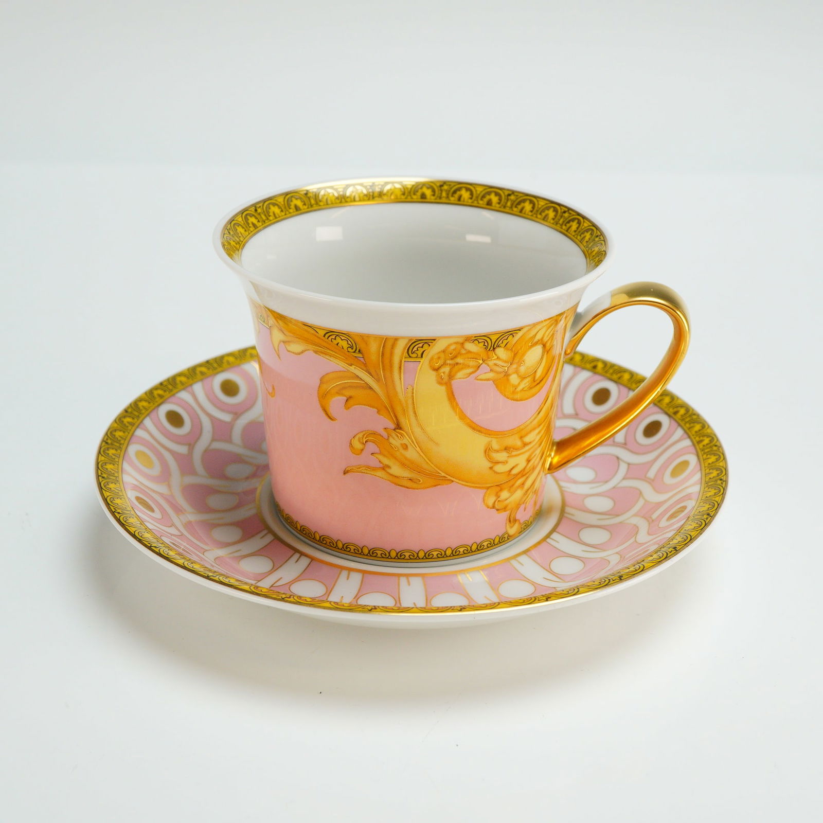 2pc Versace Rosenthal Porcelain Cup and Saucer Set, Les Reves Byzantins: The set is constructed of glazed porcelain featuring a vibrant pink ground. The cup is decorated with gold-toned baroque scrollwork and a fully gilded handle. The saucer displays a repeating geometric