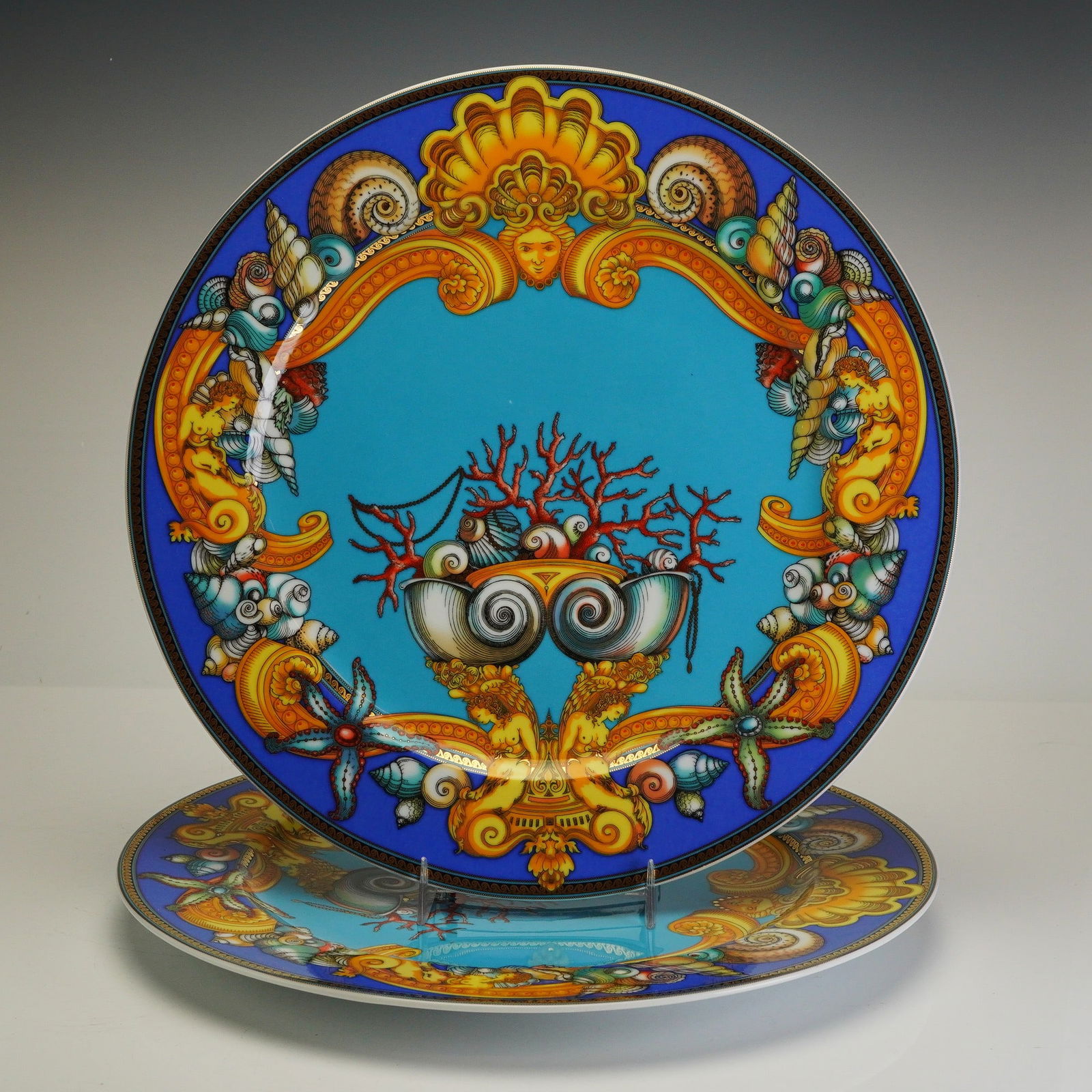Pair of Rosenthal Versace Porcelain Chargers, Les Tresors de la Mer: Each circular plate features a vibrant marine-themed design with a turquoise center field depicting coral, shells, and starfish. The center is surrounded by an ornate gold-tone baroque-style scrollwor