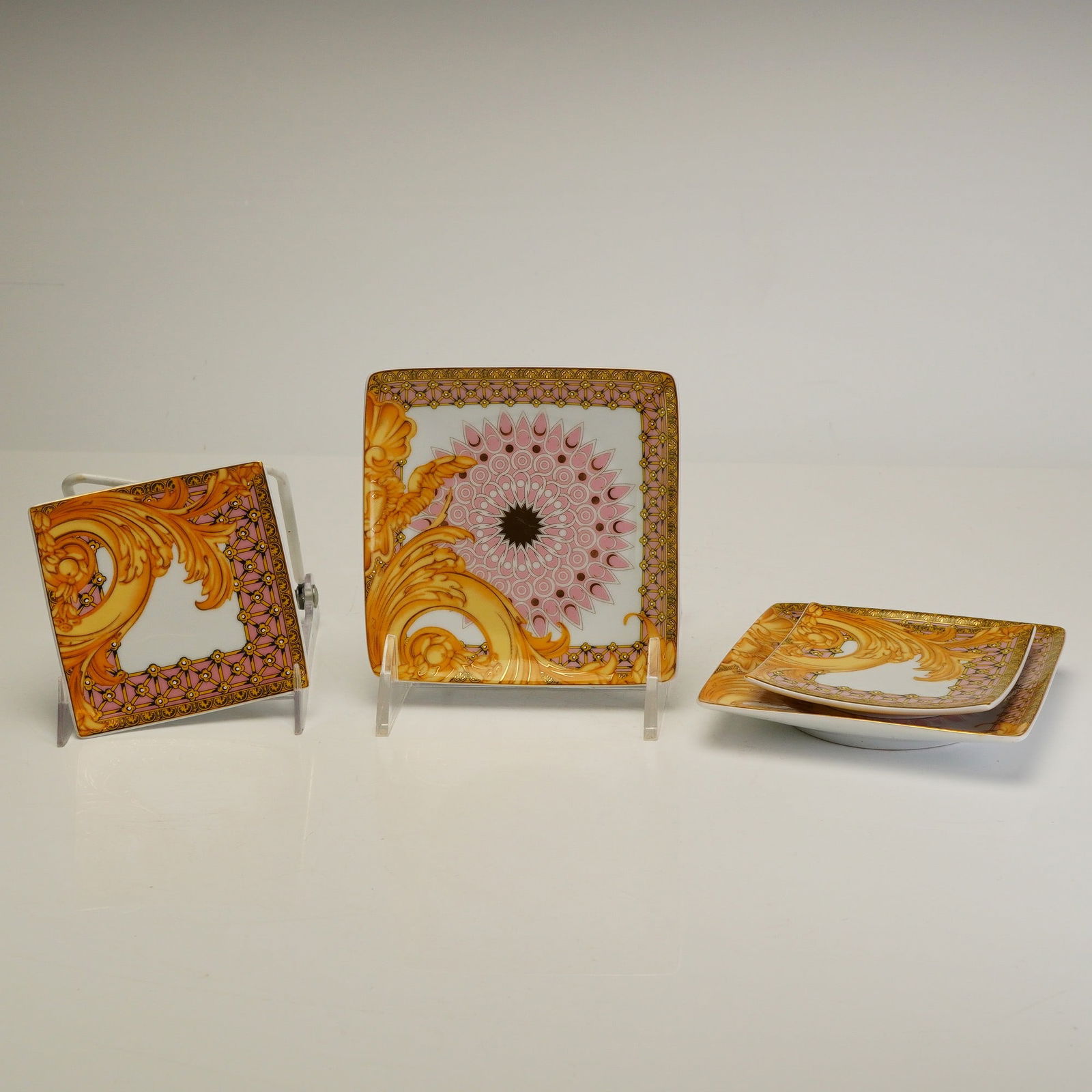4pc Versace Rosenthal Porcelain Square Dishes, Les Reves Byzantins: These contemporary pieces feature a neo-baroque design characterized by golden acanthus leaf scrollwork, a central pink and white geometric sunburst motif, and a pink latticework border with gilded ac