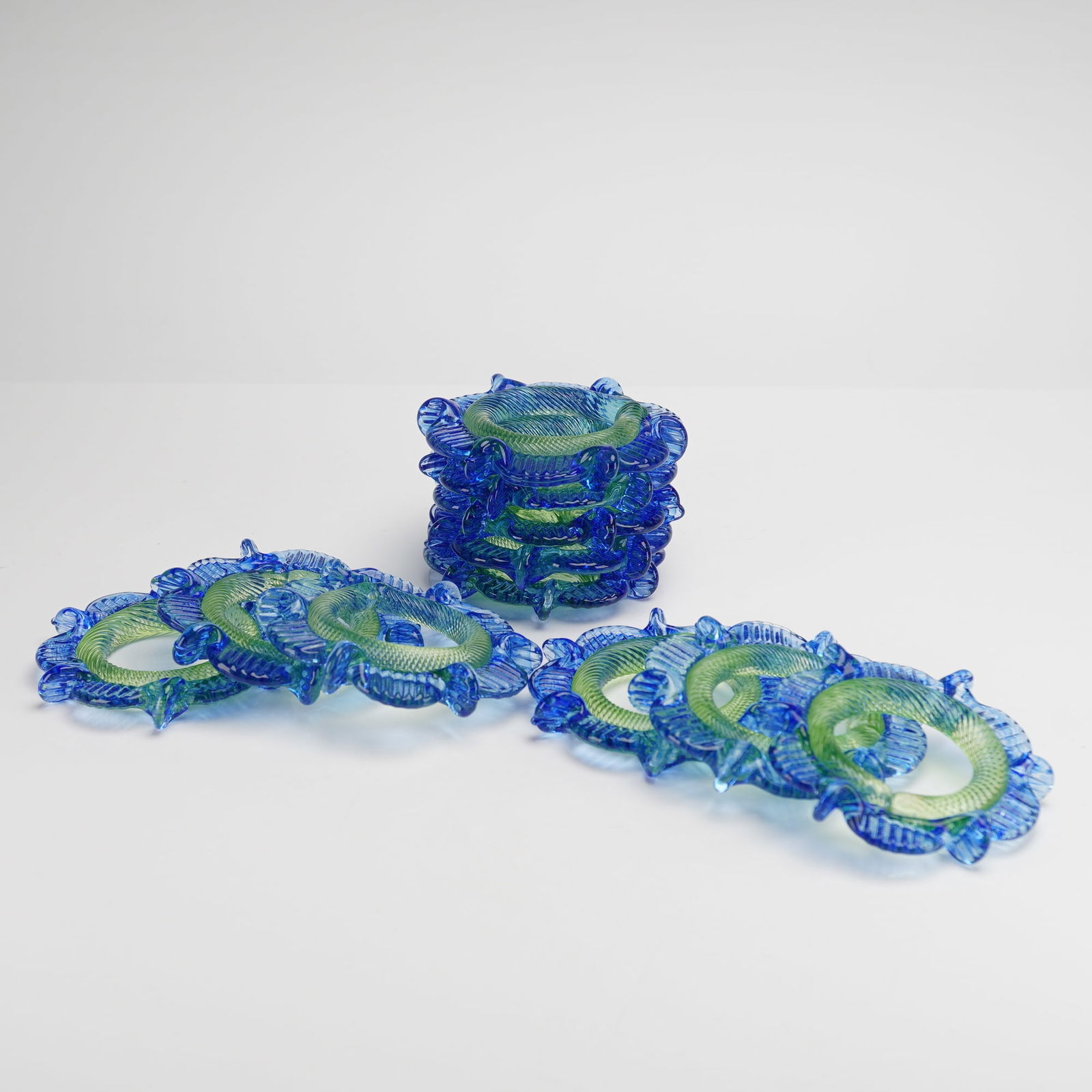 11pc Art Glass Floral Blue and Green Napkin Rings: A collection of eleven Mid-Century Modern Glass Floral Rosettes napkin rings. Each ring is constructed from multicolored art glass. The central ring core utilizes a twisted green and chartreuse cane