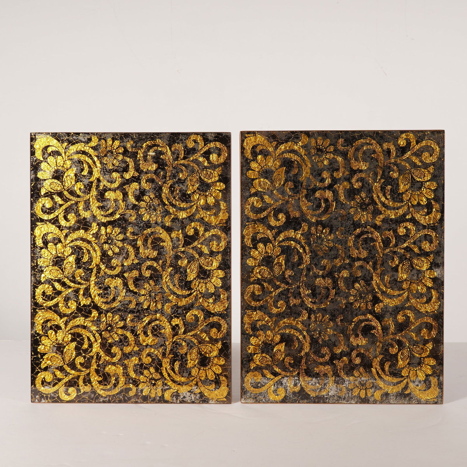 2pc Nomi K Black and Gold Glass Lace Motif Mirror Placemats: Two rectangular mirror placemats by Nomi K featuring gold leaf detailing in a lace motif of floral scrollwork and foliate patterns. The design is set against a dark, mottled background with a crackled