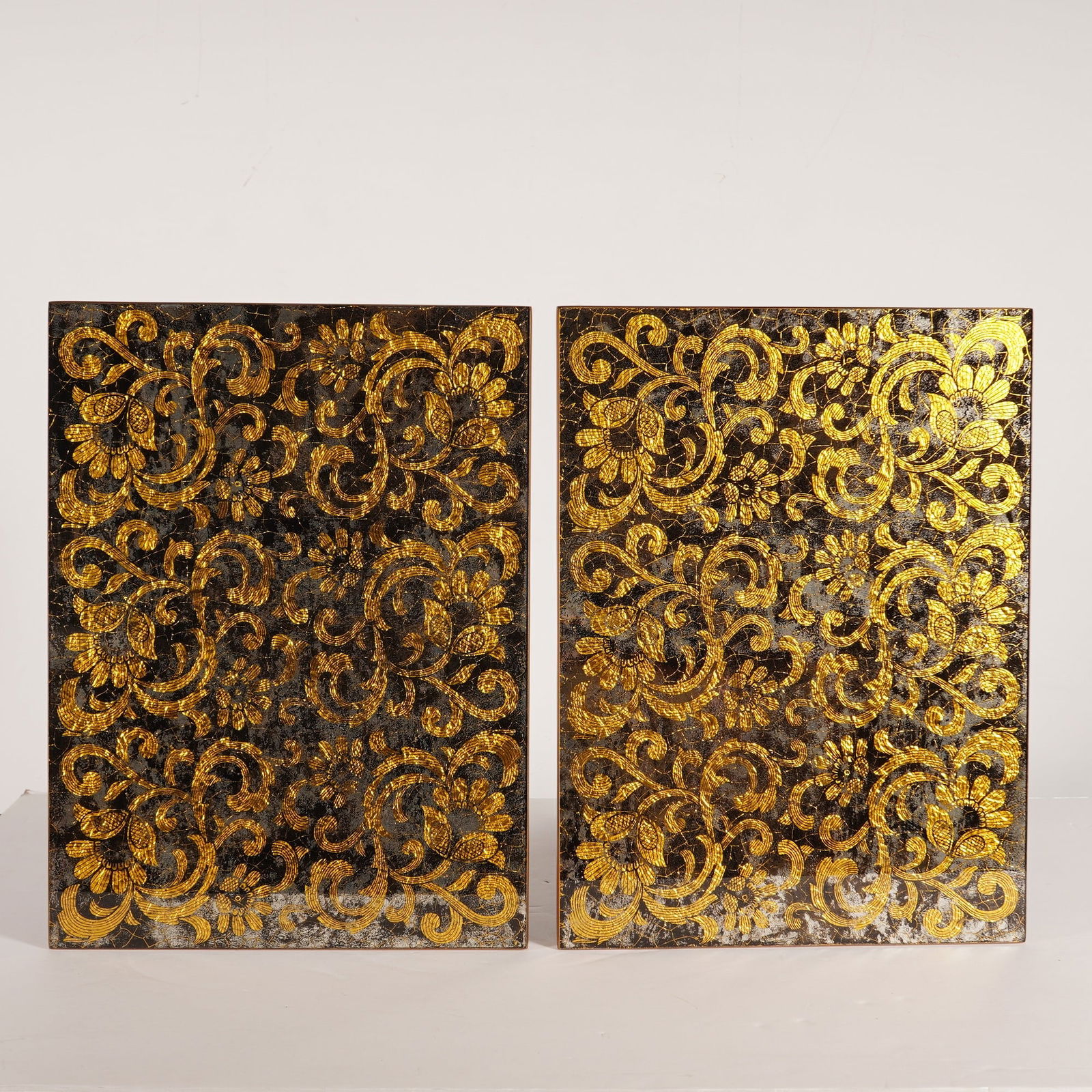2pc Nomi K Black and Gold Glass Lace Motif Mirror Placemats: A pair of Nomi K black and gold glass lace motif mirror placemats featuring gold leaf detailing and wood backing. Each rectangular panel displays a gold scrolling floral and foliate design with textur