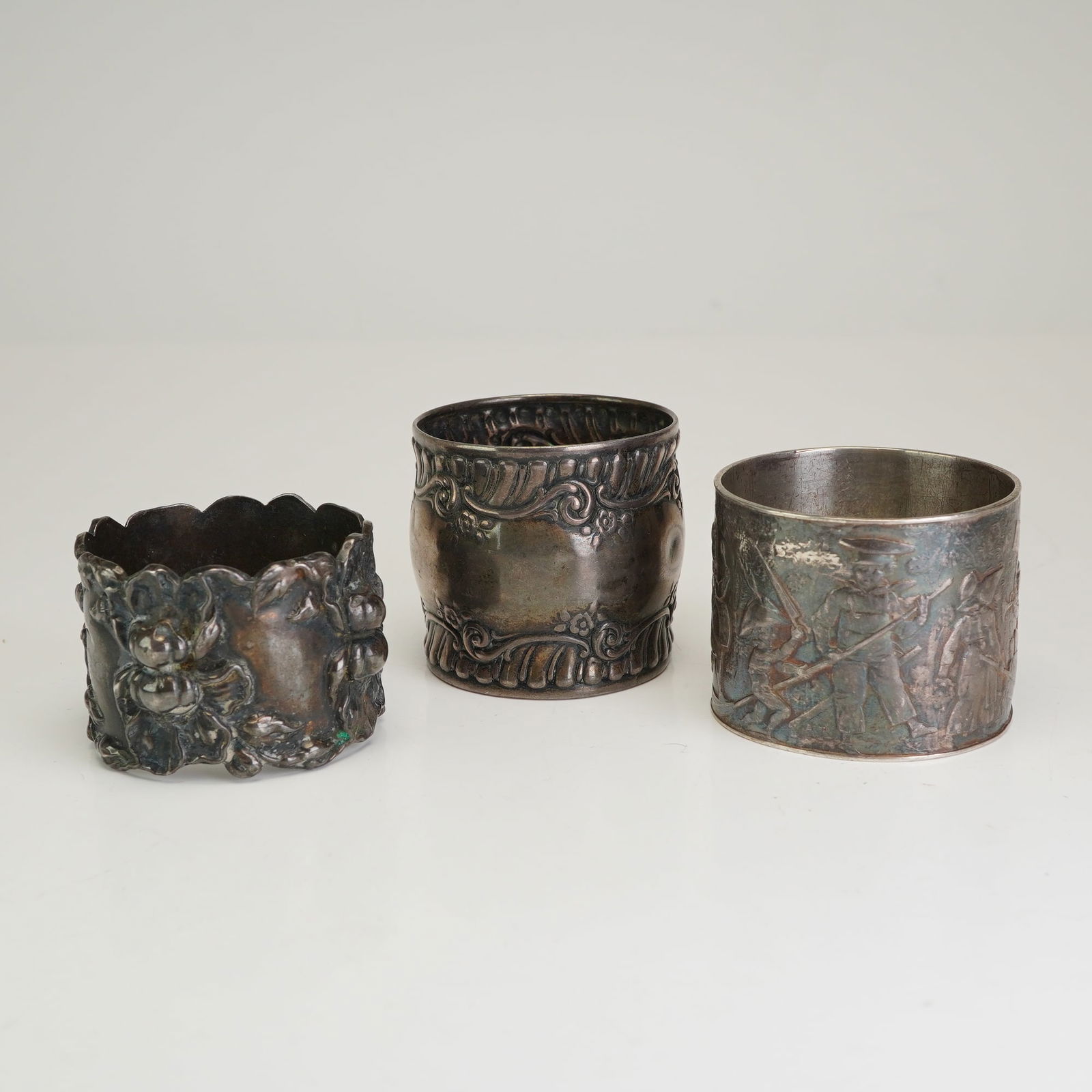 Three Monogrammed Sterling Silver Napkin Rings: This lot consists of three sterling silver napkin rings in various decorative styles. The first ring features a heavy repousse floral motif with a scalloped rim. The second ring is cylindrical with a