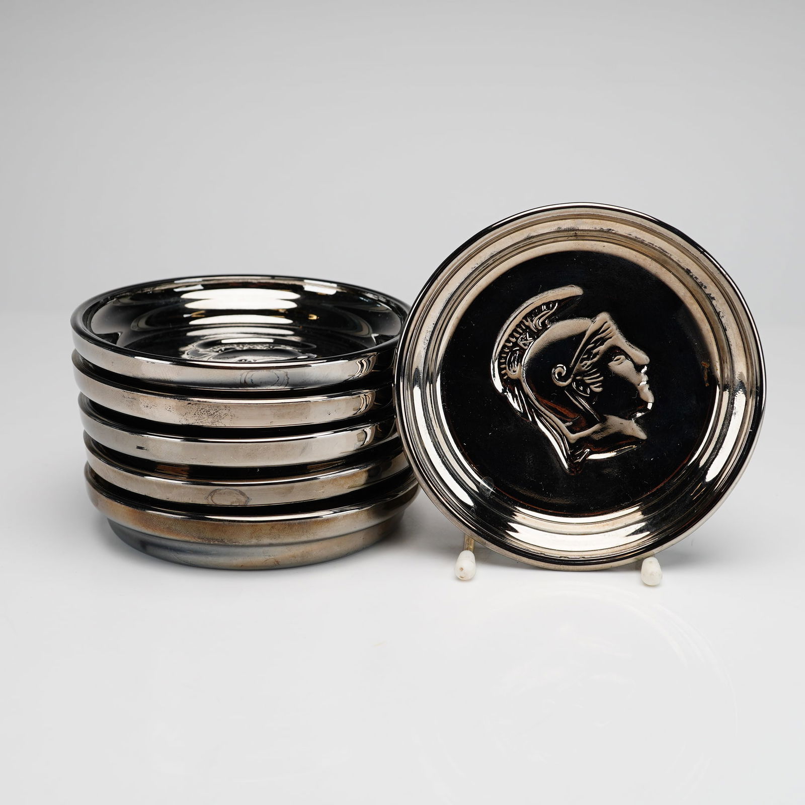 Set of Six Roman Bust Silver-Plated Coasters: Coasters feature a silver-plated finish over a glass body, consistent with mercury glass construction. Each piece is decorated with a central relief depicting a classical Roman bust in profile wearing