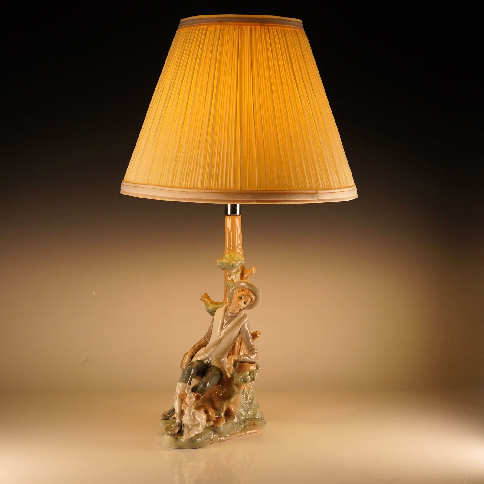 Lladro Porcelain Figural Table Lamp, Shepherd 1004659: A fine glazed porcelain table lamp featuring a hand-painted figurine depicting a young boy in a hat leaning against a tree trunk with a small dog resting at his feet, finished in a traditional pastel