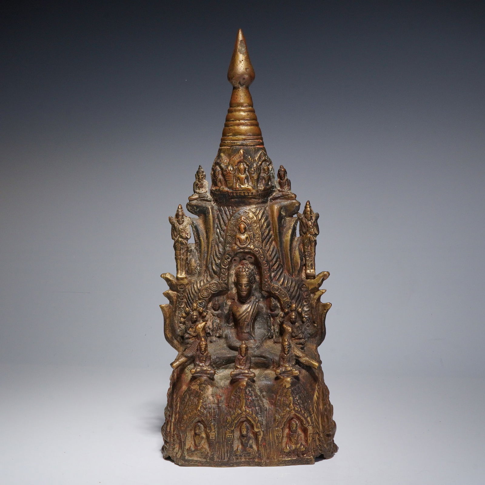 Southeast Asian Cast Bronze Buddhist Shrine: The tapering architectural form rising from a flared base to a stupa-form finial, the front centered by a seated figure identified as Gautama Buddha in meditation posture within a tiered mandorla, sur