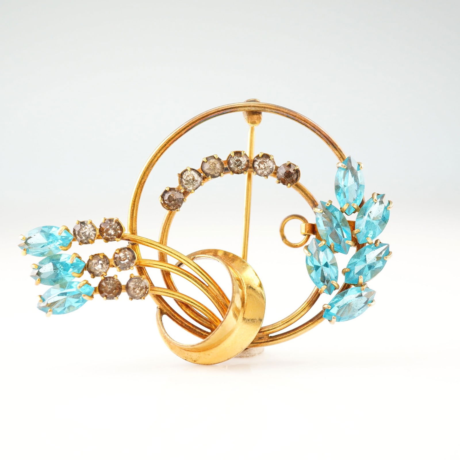 Carl-Art 12K Gold Filled Blue Floral Brooch: This mid-century brooch by Carl-Art Inc. is crafted in 12K gold-filled metal and features a stylized floral and foliate spray set within concentric circular frames. The design is accented with nine ma