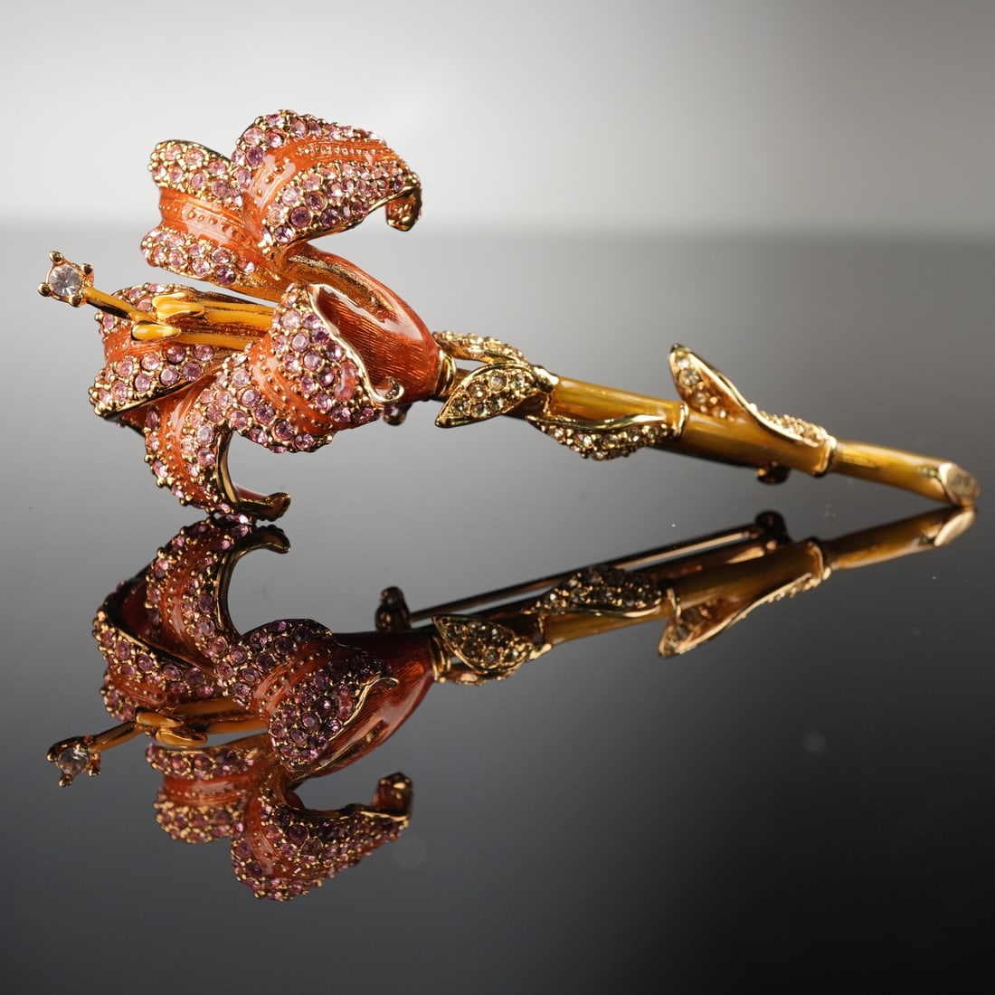 Nolan Miller Style Orange Enamel and Pink Rhinestone Lily Brooch: A Nolan Miller style lily brooch, fashioned from gold-toned metal. The piece features lustrous orange enamel on the petals and yellow enamel on the interior of the flower and part of the stem. The pet