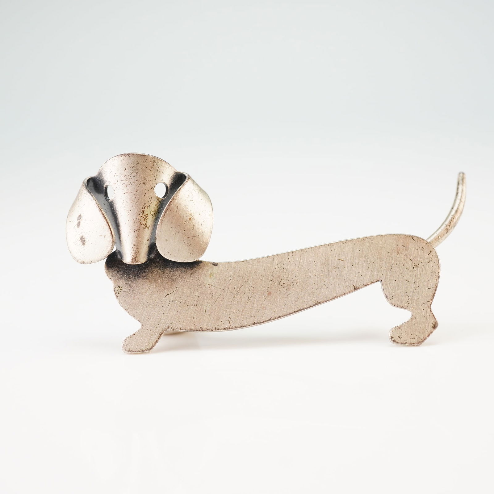 Sterling Silver Dachshund Brooch: This sterling silver brooch depicts a stylized dachshund dog in a minimalist Mid-Century Modern style. The head section is folded to create a three-dimensional effect with drooping ears and pierced ci