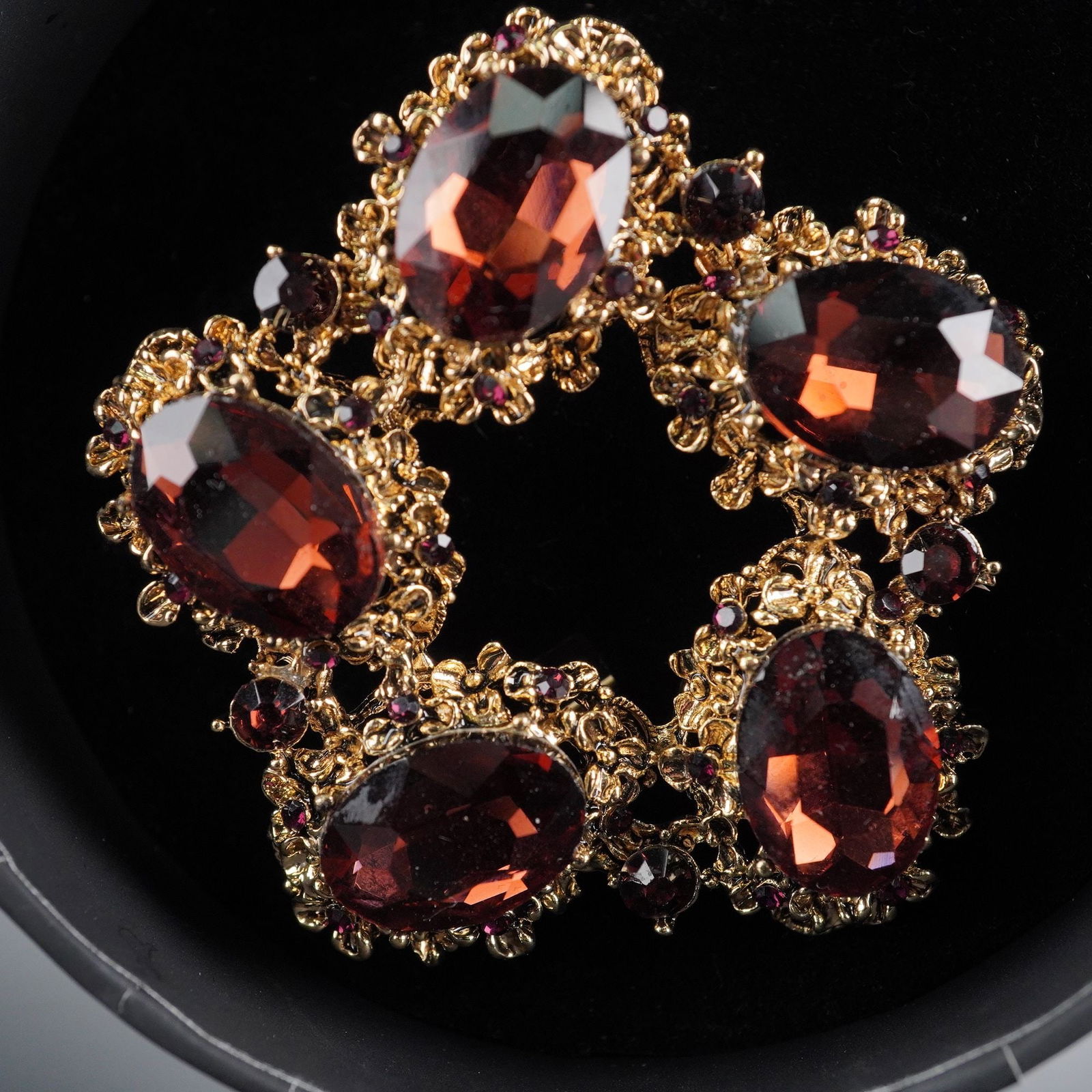 A Natasha Flower Shaped Burgundy Costume Brooch: A contemporary five petal flower costume brooch, constructed from gold-toned metal. The brooch is set with five large oval-cut, faceted dark burgundy colored stones, accented by numerous smaller round