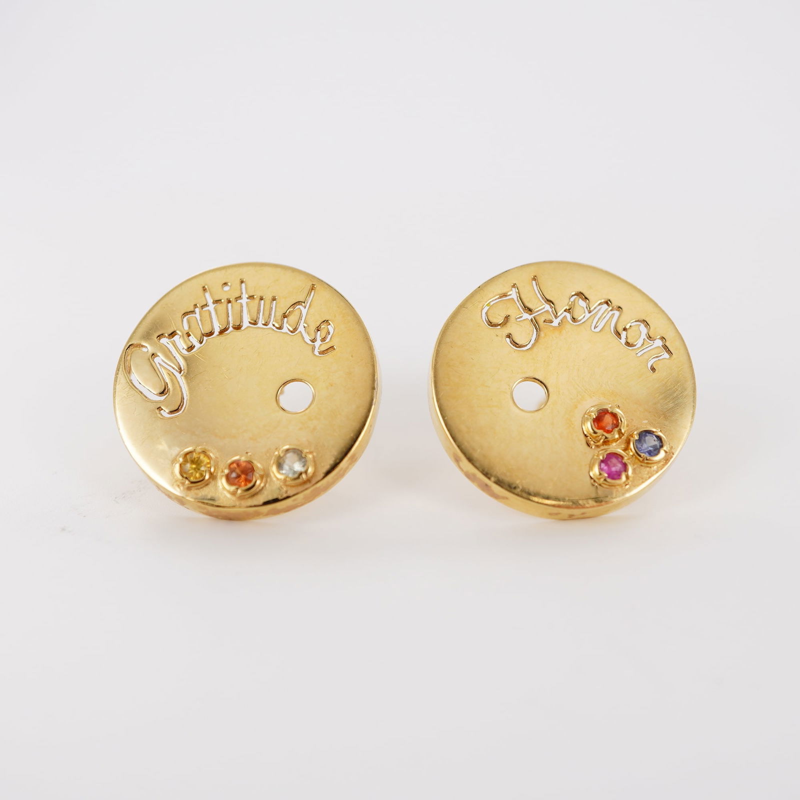 Pair of 18K Gold Earrings with Gemstones, Engraved Gratitude and Honor: A pair of 18K gold circular stud earrings script engraved "Gratitude" and "Honor." On their lower parts, three small, round, bezel-set yellow, orange, pink,/red, purple, and light blue tones tested ge