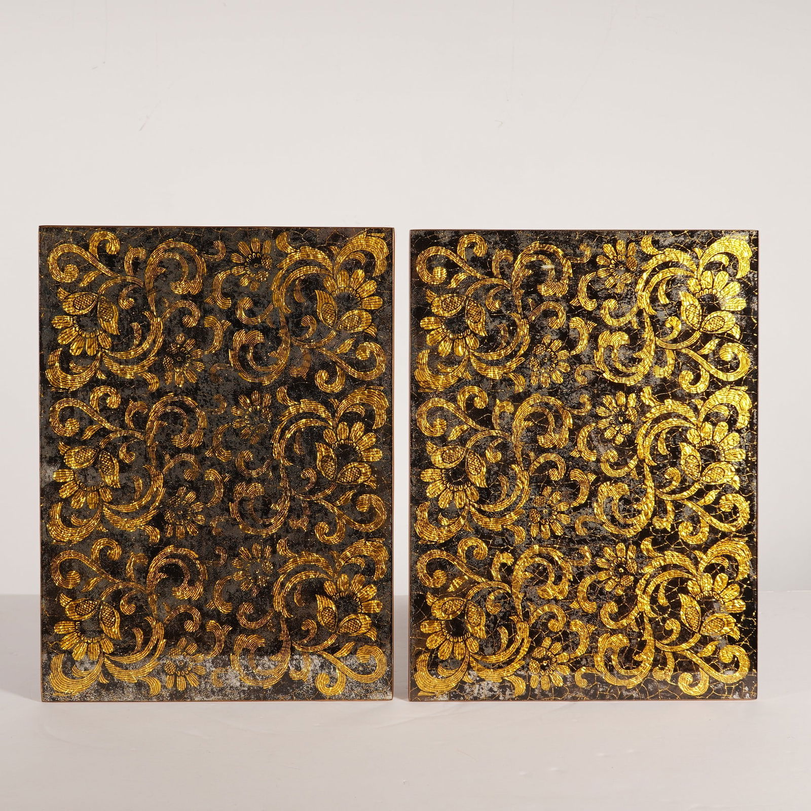 2pc Nomi K Black and Gold Glass Lace Motif Mirror Placemats: A pair of Nomi K black and gold glass lace motif mirror placemats featuring gold leaf detailing and wood backing. The rectangular panels display an ornate, textured gold-tone floral and scrollwork pat