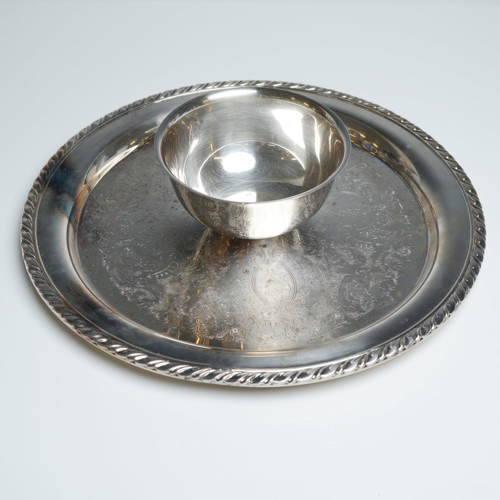 Oneida Silverplated Park Lane Chip and Dip Serving Platter: The piece features a circular tray with an intricately etched floral and foliate center, framed by a raised rim with a decorative rope-twist border. The attached center bowl is of a simple, rounded fo