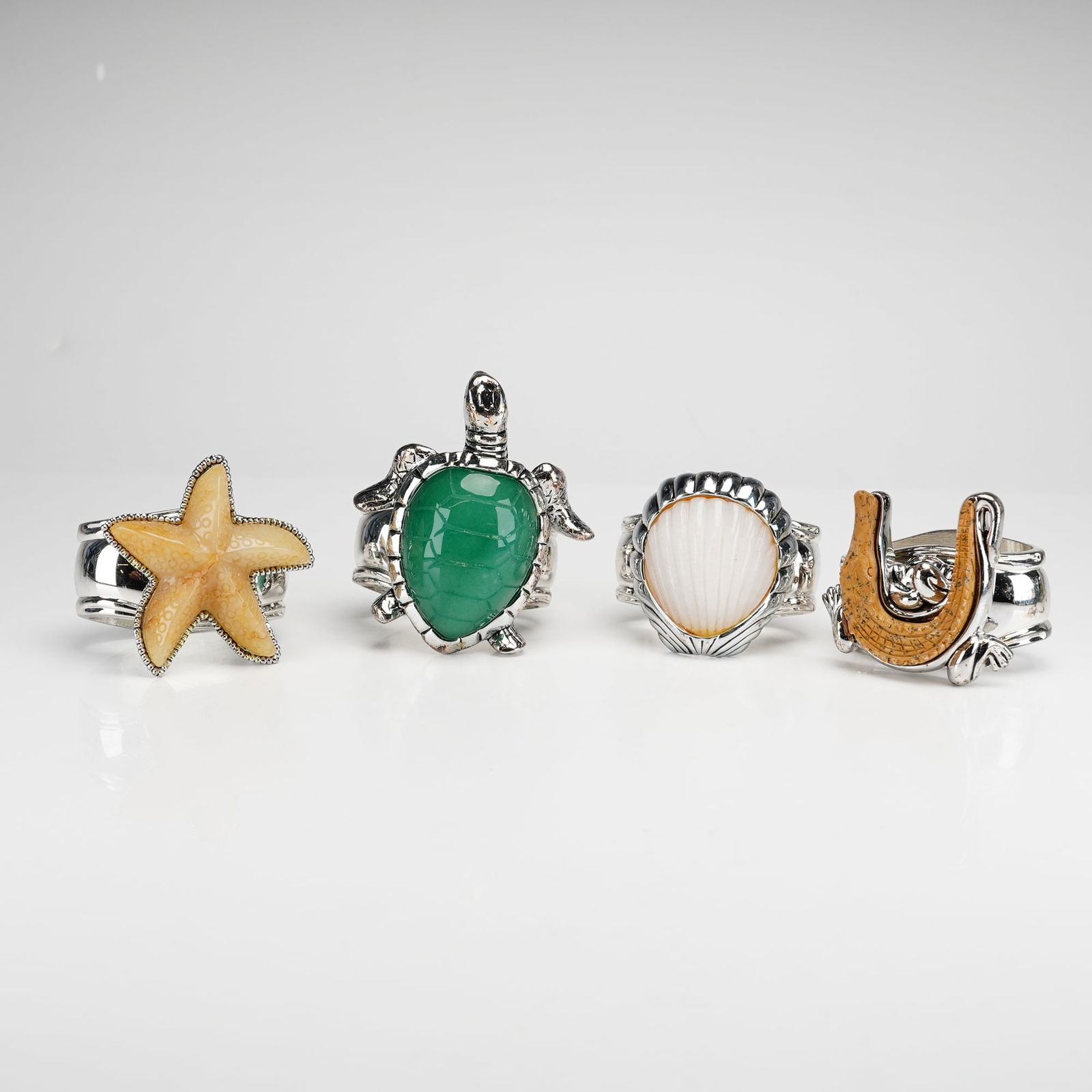 4pc Hans Turnwald Napkin Rings: A set of four napkin rings by Hans Turnwald, featuring various nature-themed motifs. The collection includes a starfish set with a textured yellow stone, a sea turtle with a green cabochon shell, a sc