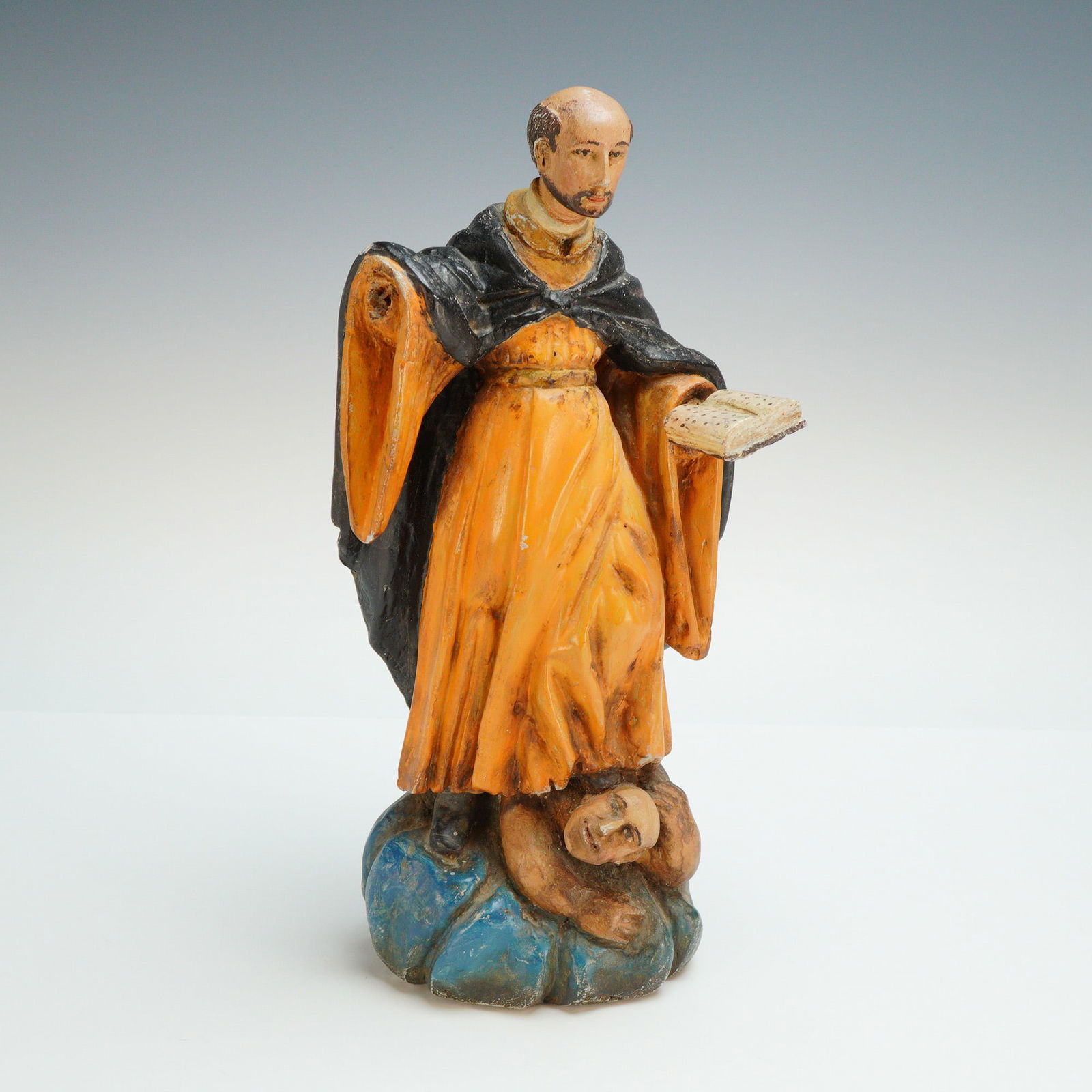 Antique Polychromed Wooden Santos Sculpture of Saint Vincent: The figure is rendered in clerical attire consisting of an orange-yellow tunic beneath a black hooded cape. He is depicted holding an open book in his left hand and stands atop a blue cloud-form base.