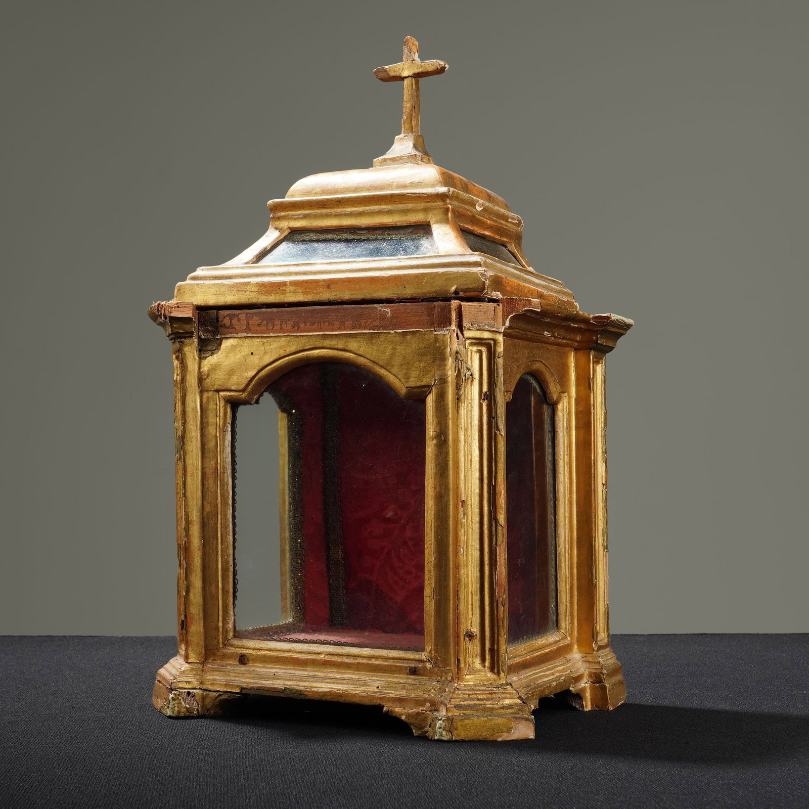Giltwood Display Reliquary Shrine: This carved giltwood reliquary shrine features a tiered architectural frame with arched display panels on three sides and a roof topped with a cross finial. The interior is lined with red patterned fa