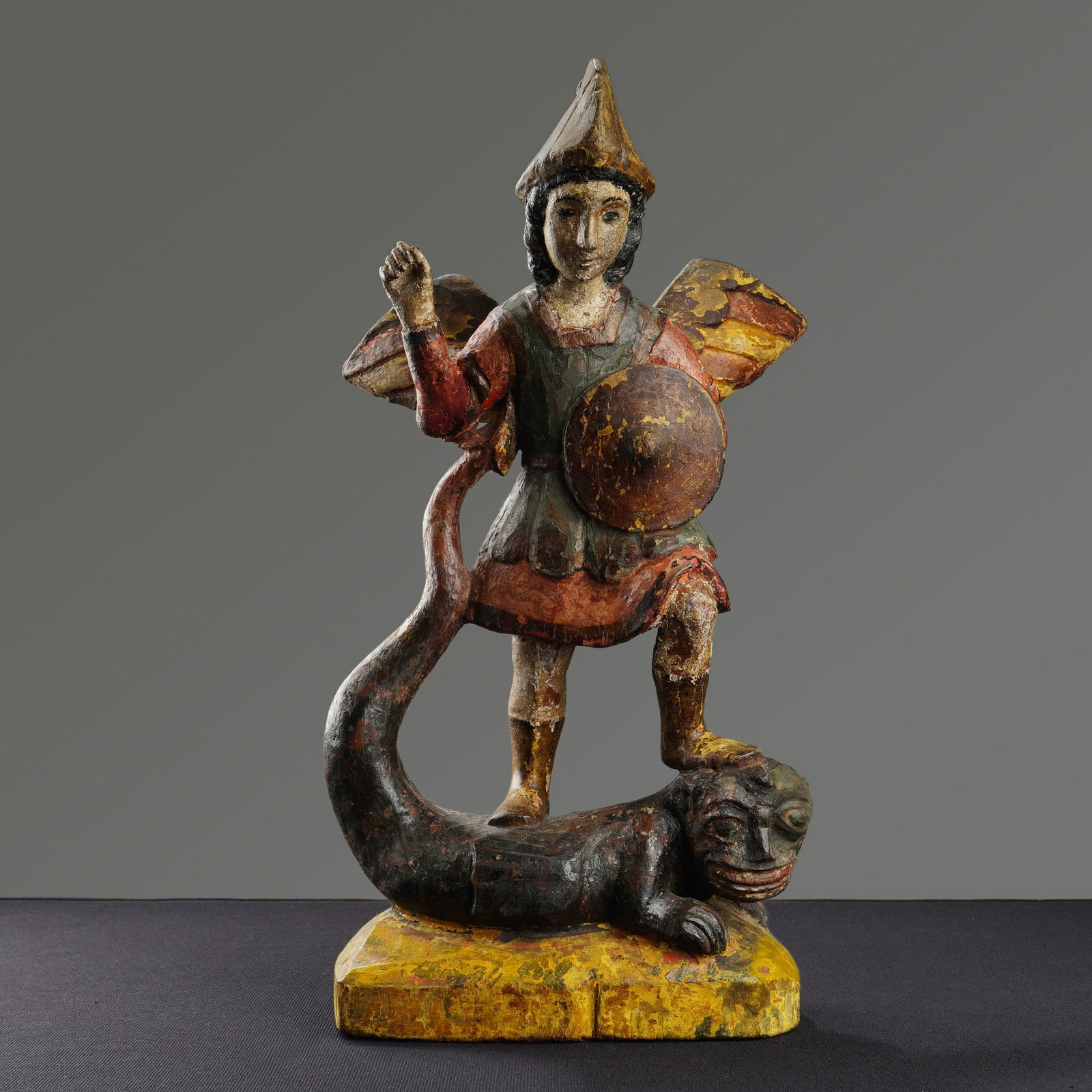 Spanish Colonial Saint Michael Archangel Wood Figure: A Spanish Colonial Mexican polychrome carved wood statue of Saint Michael the Archangel standing over a defeated Satan. This religious folk-art figure is depicted in a triumphant pose, treading upon a