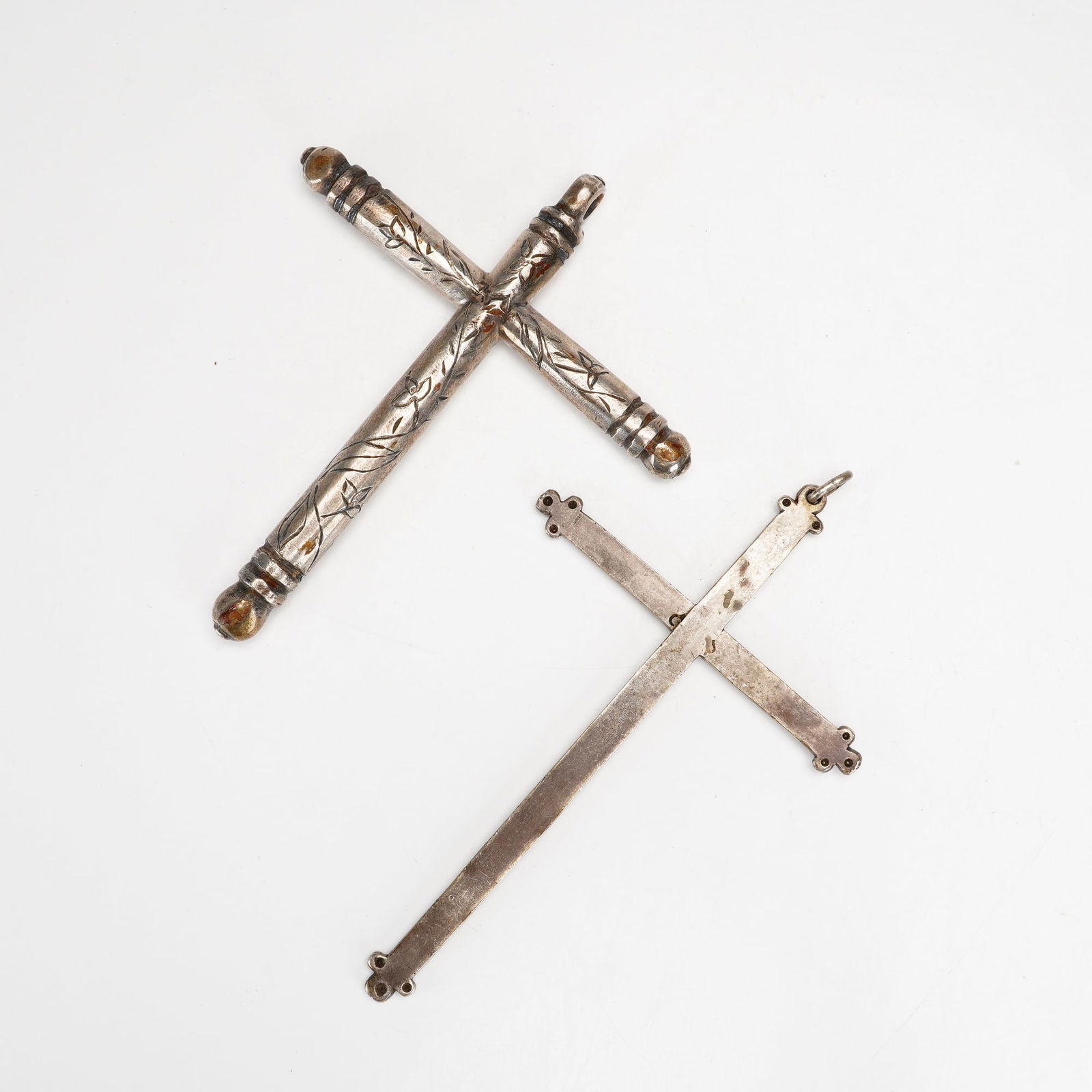 2pc Metal Etched Floral Crucifix Pendants: This lot comprises two etched silver-toned metal crucifix pendants: one with a three-dimensional tubular form featuring bulbous, tiered finials and hand-engraved floral and foliate patterns across its