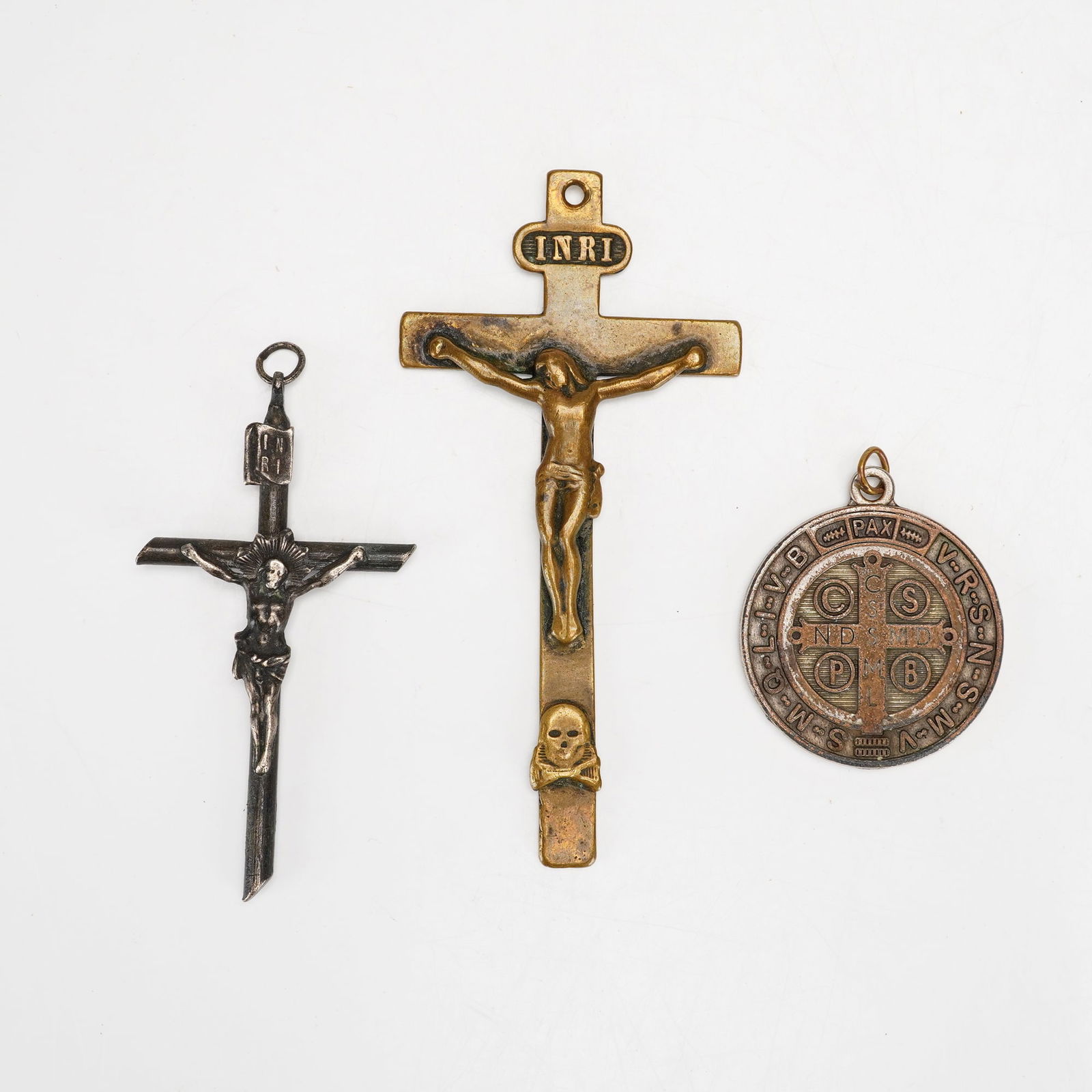 3pc Crucifix and St. Benedict Medal Religious Pendants: A collection of three religious pendants comprising two crucifixes and one Saint Benedict medal. The first crucifix is constructed of a dark, silver-toned metal featuring a corpus and a plaque at the