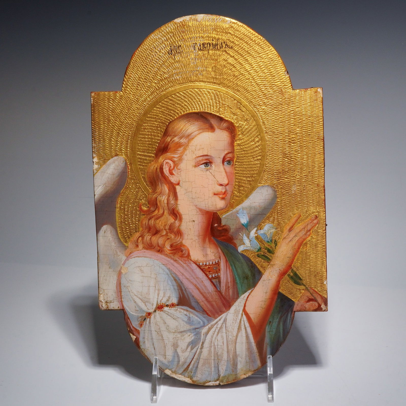 Russian Orthodox Icon of Archangel Gabriel: This Russian Orthodox depiction of the Archangel Gabriel is executed in tempera on a shaped wood panel with a gilded carved background. The figure is shown from the chest up, holding a white lily agai