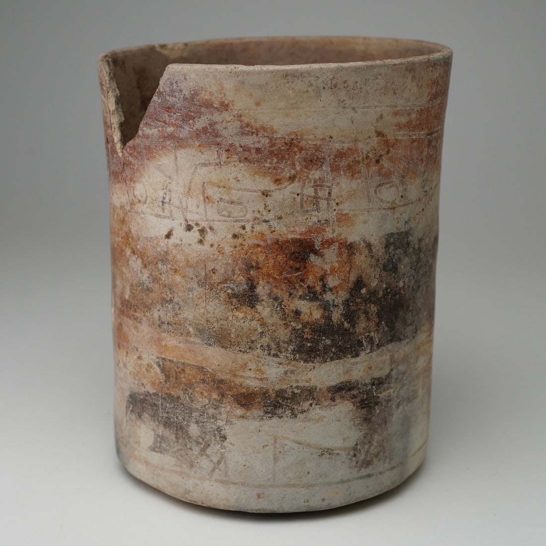 Pre-Columbian Cylindrical Earthenware Vessel with Carvings: This earthenware vessel features a cylindrical form and displays a weathered surface with varied earthy hues. The exterior is decorated with incised carvings, consisting of linear and geometric patter