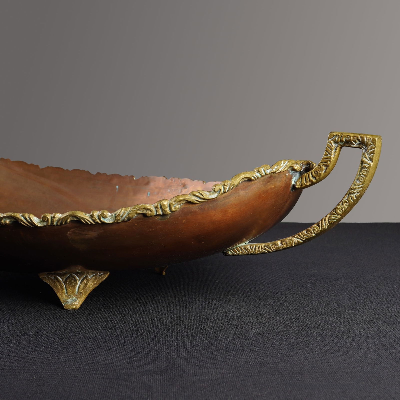Mexican Copper and Brass Footed Centerpiece Bowl: A vintage footed centerpiece bowl of elongated oval form, featuring a copper body with ornate cast brass mounts. The design includes a scrolling foliate border along the rim and two stylized side hand