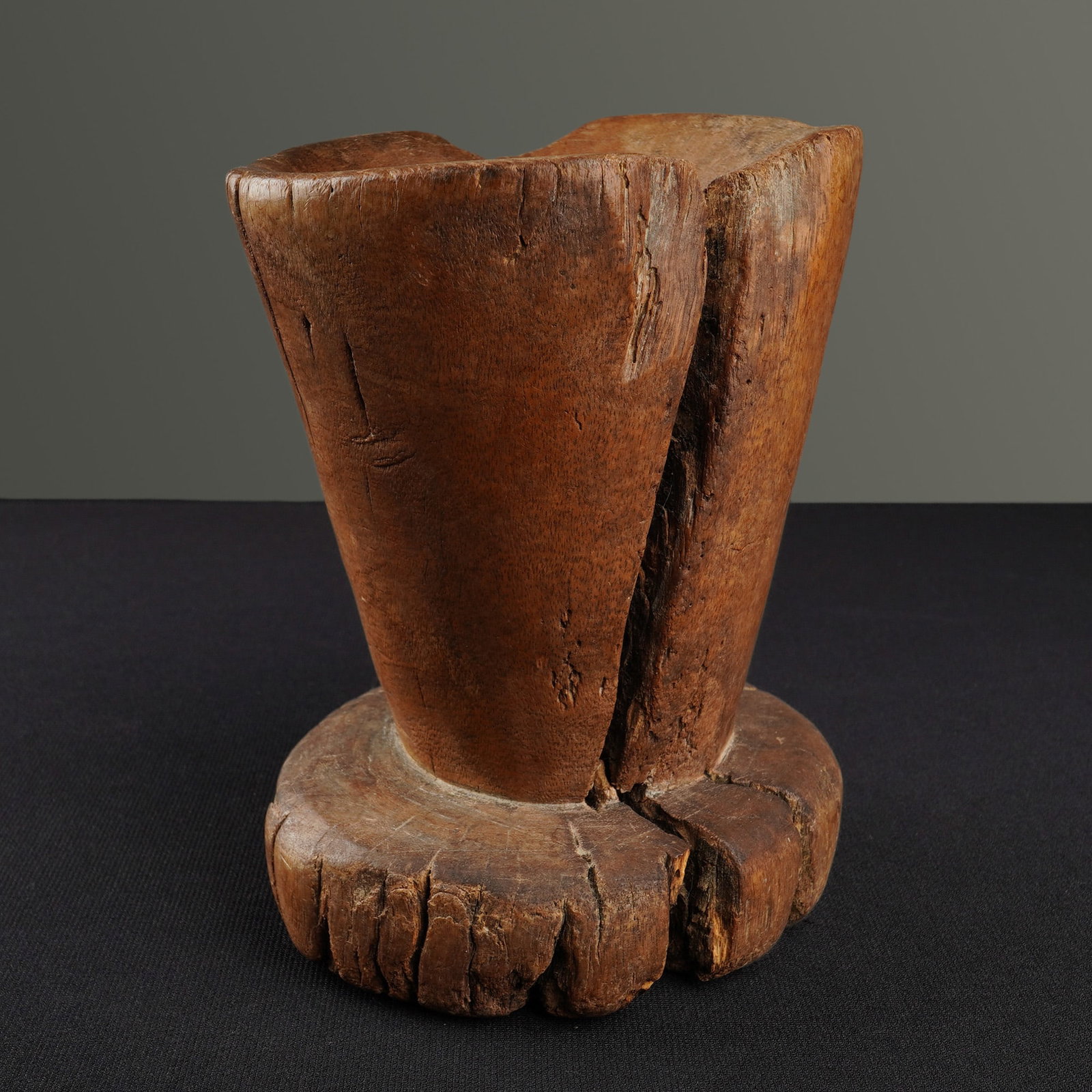 Hand Carved Wooden Mortar: This carved wooden mortar is fashioned from a single block of dense hardwood. The vessel features a thick, circular pedestal base supporting a flared, hollowed-out conical body. The exterior surface